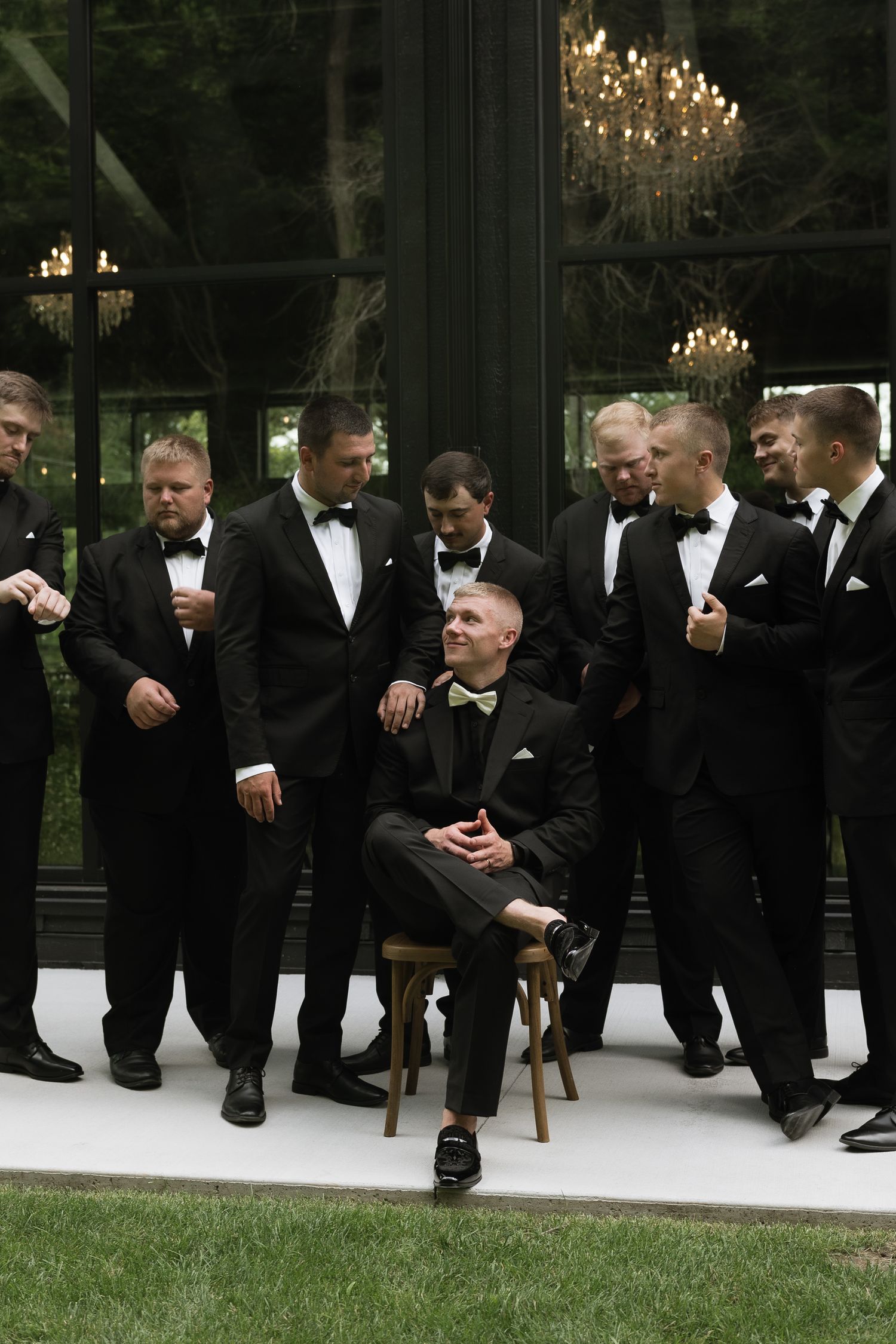 Groom seated on chair surrounded by groomsmen in black tuxedos outside glass venue with chandelier.