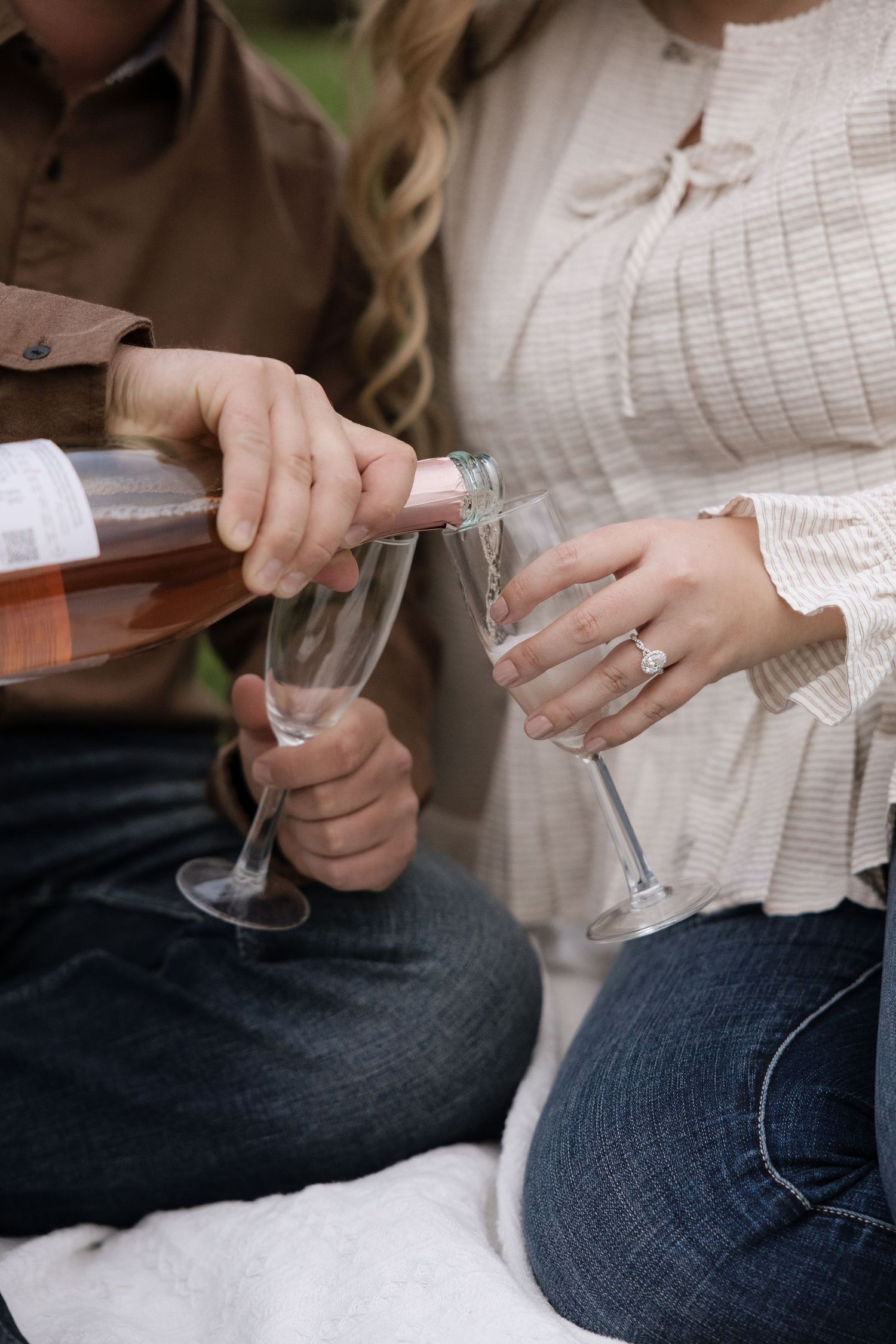 Couple celebrating engagement with rose champagne pour into flutes, woman displaying diamond ring, outdoor picnic setting.