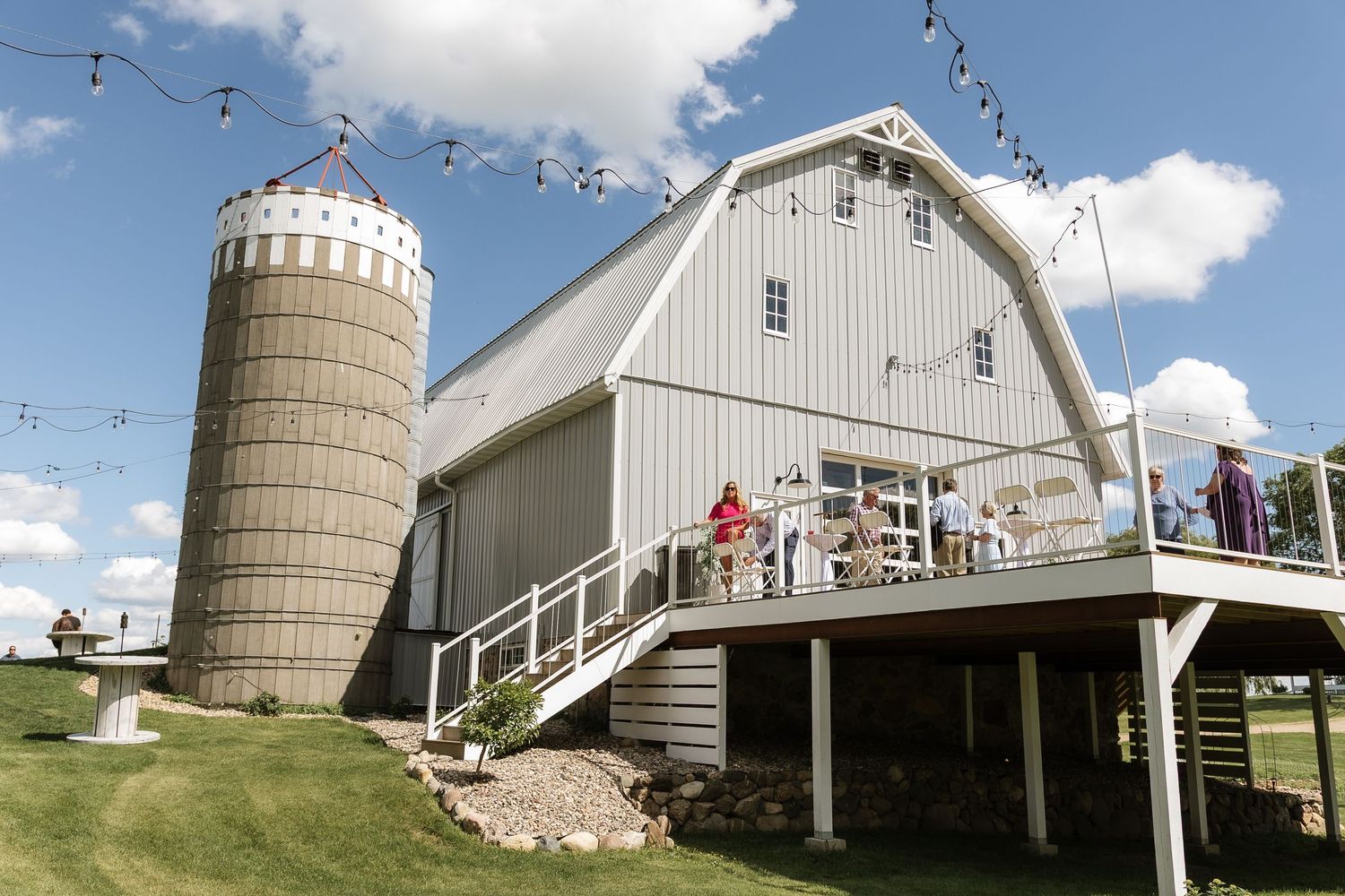 Rustic gray barn wedding venue with white silo, string lights, and guests on wooden deck under blue sky.