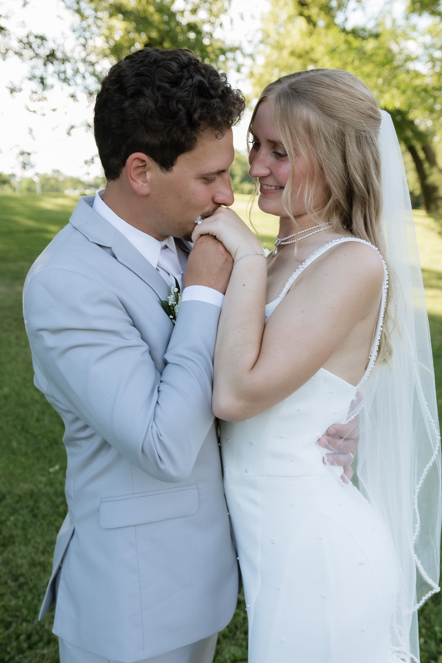 Groom in gray suit kissing bride's hand outdoors, with bride in white wedding dress and veil smiling softly.