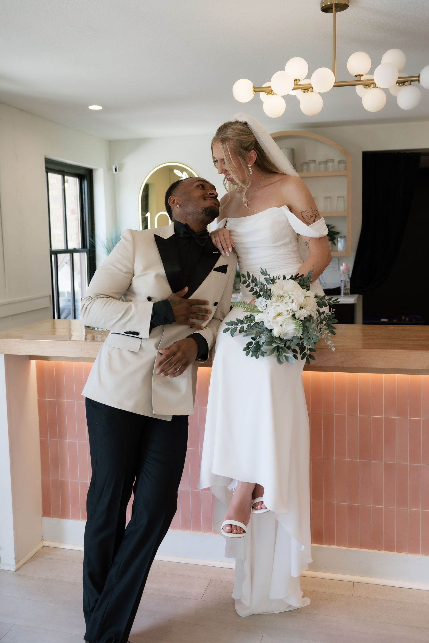 Interracial couple in wedding attire sharing a romantic moment, bride holding white bouquet, groom in cream blazer, pink tiled bar backdrop.