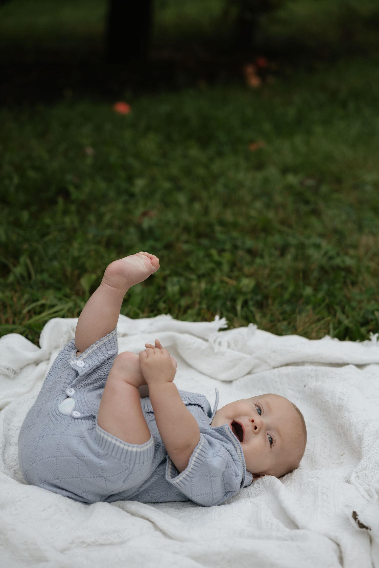 Cute baby in gray romper lying on white blanket on green grass, smiling and holding feet up in the air.