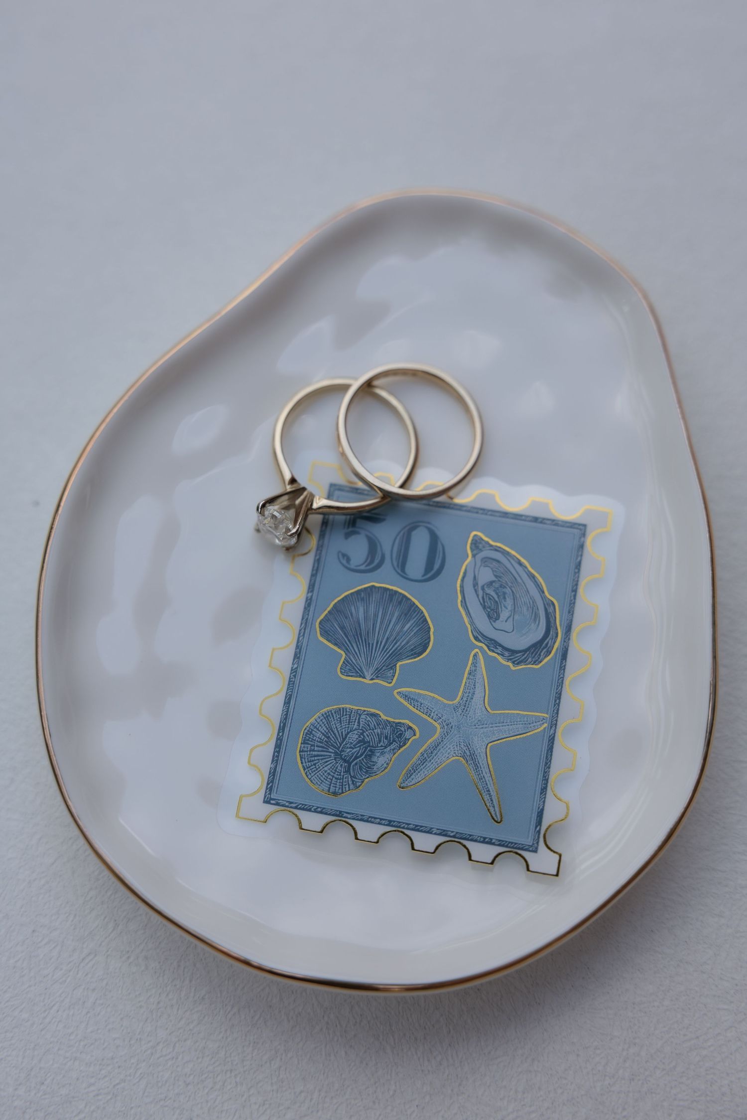 Gold wedding rings and engagement ring on a white ceramic dish with a blue seashell stamp card.