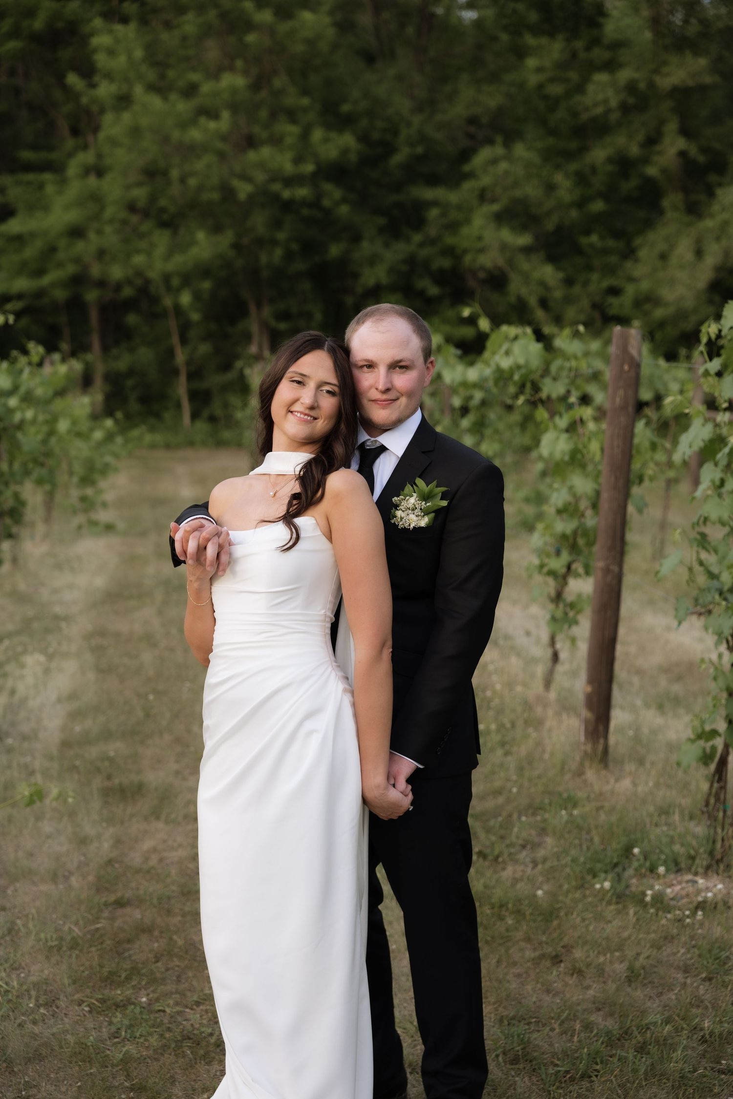 Bride and groom pose together in a vineyard, groom embracing bride from behind among green vines.