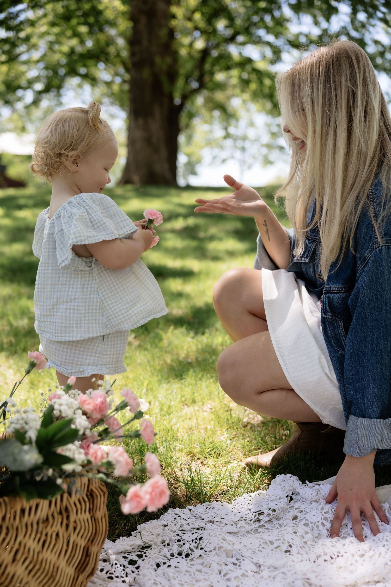 Blonde mother crouches down smiling at her toddler daughter holding a flower during park picnic.