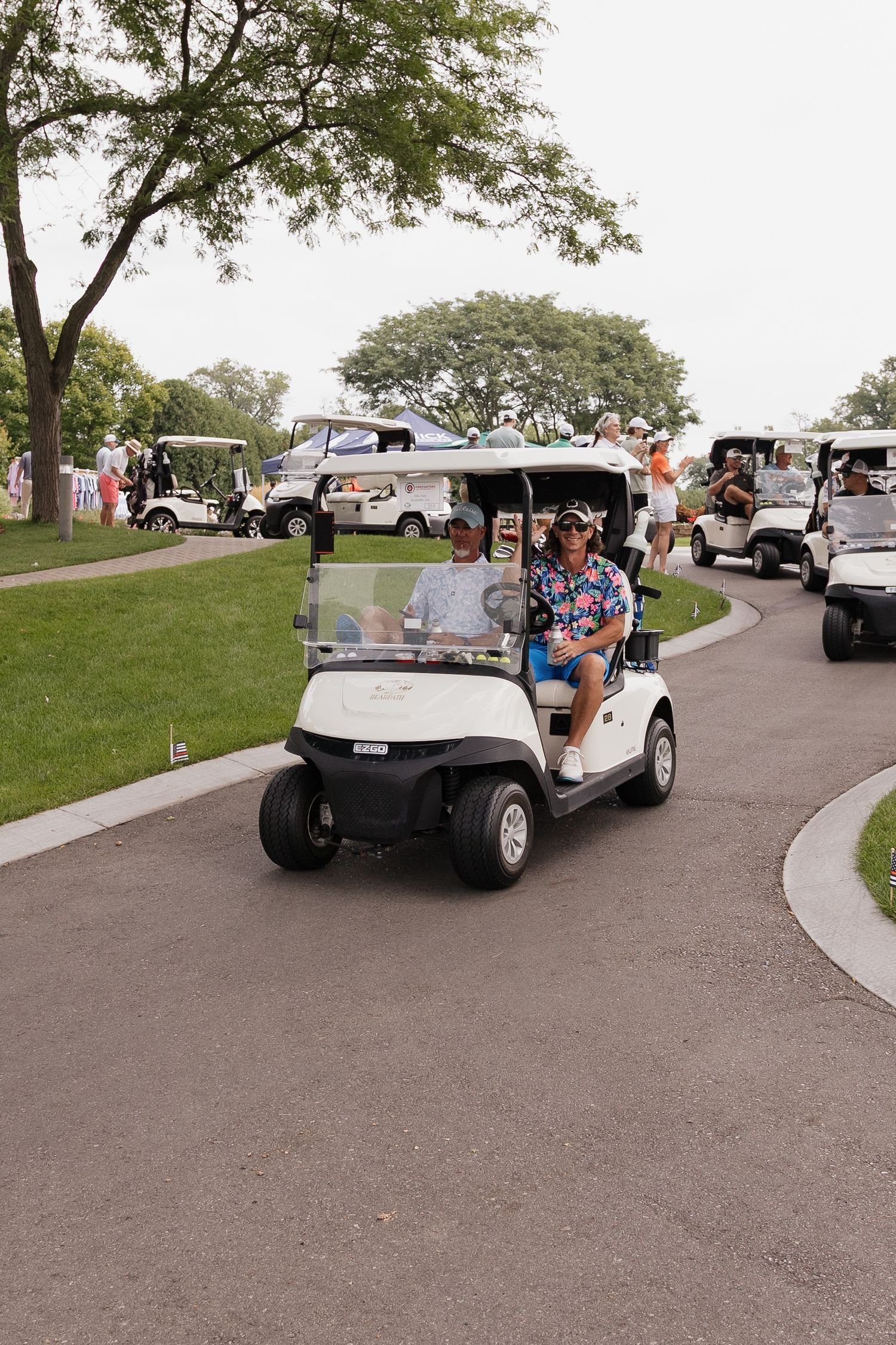 Two golfers ride a white golf cart on a sunny course path during a golf tournament outing.