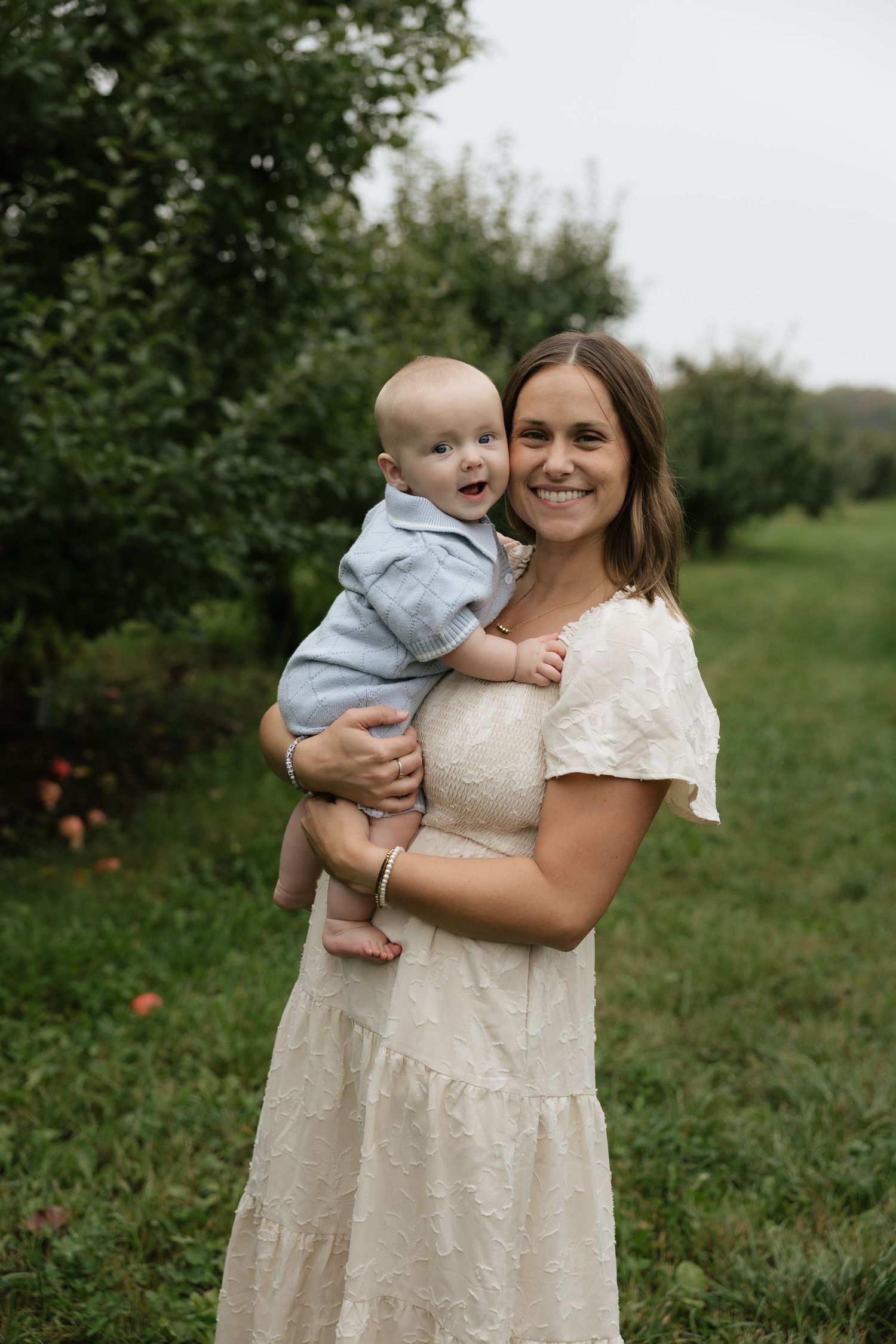 Smiling mother in white lace dress holding happy baby boy in blue outfit in a lush green orchard.