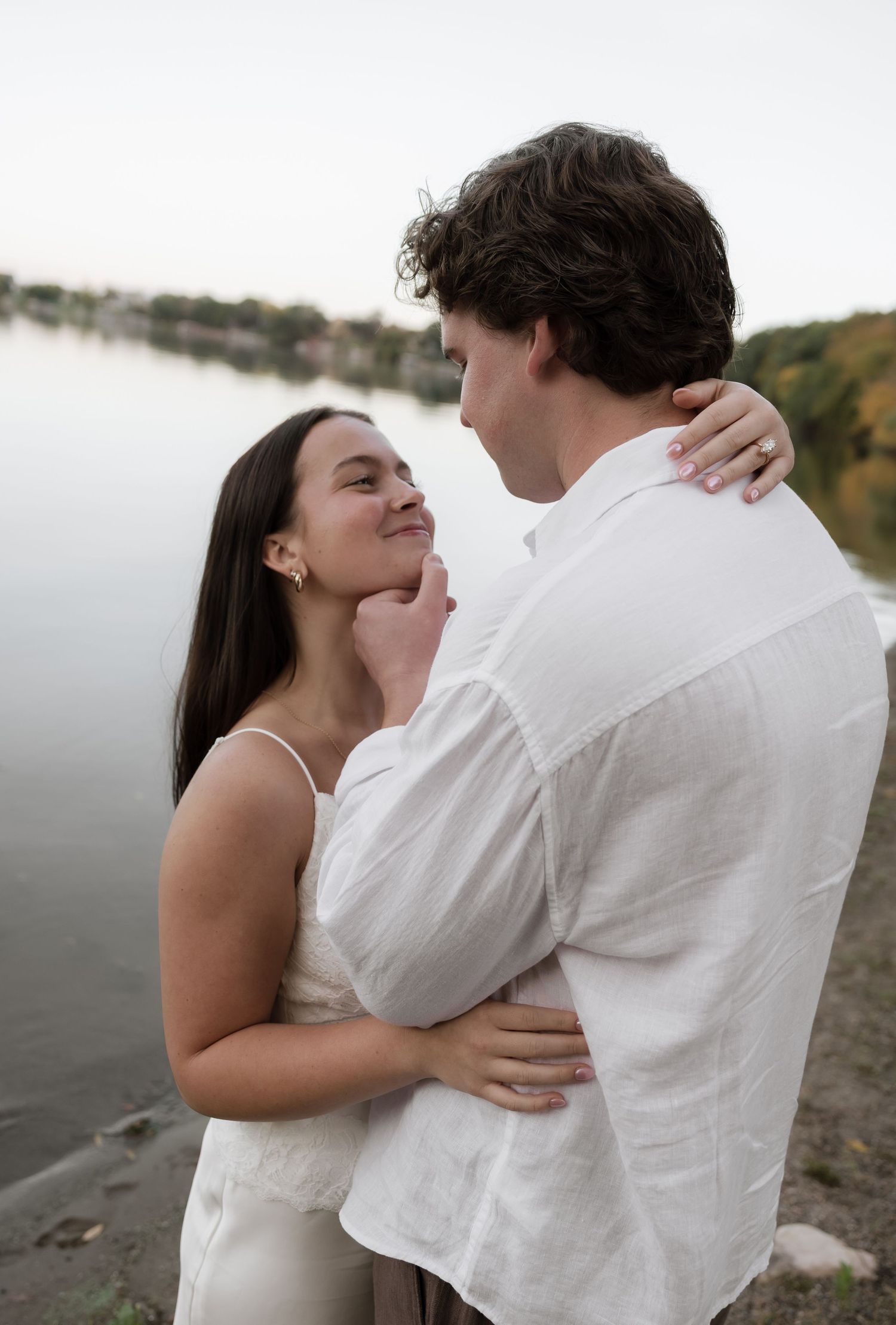 Couple embracing tenderly by a lake, woman in white dress smiling up at man in white linen shirt during engagement session.