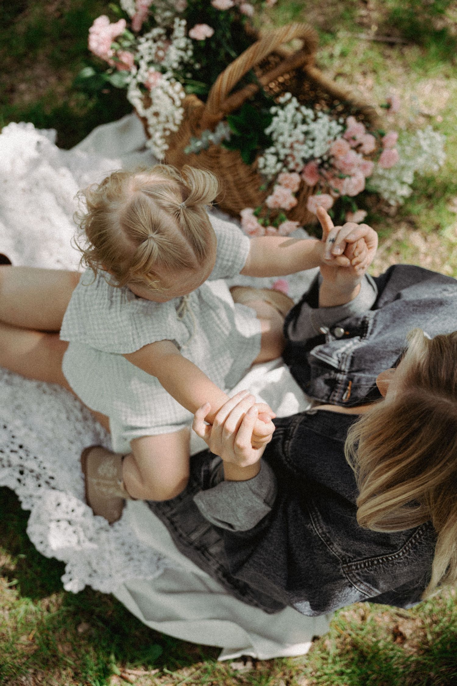 Overhead view of mother and toddler holding hands on white blanket at outdoor picnic with floral basket.