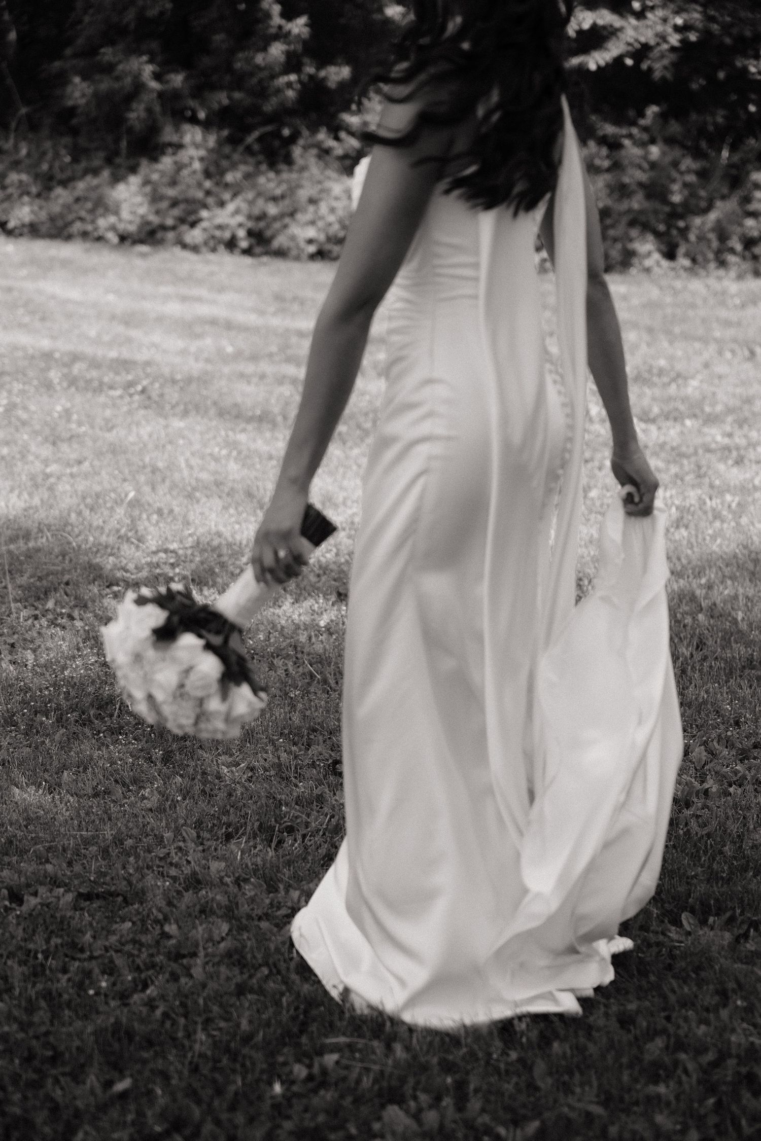 Black and white photo of bride in flowing white gown holding bouquet while walking on grass.