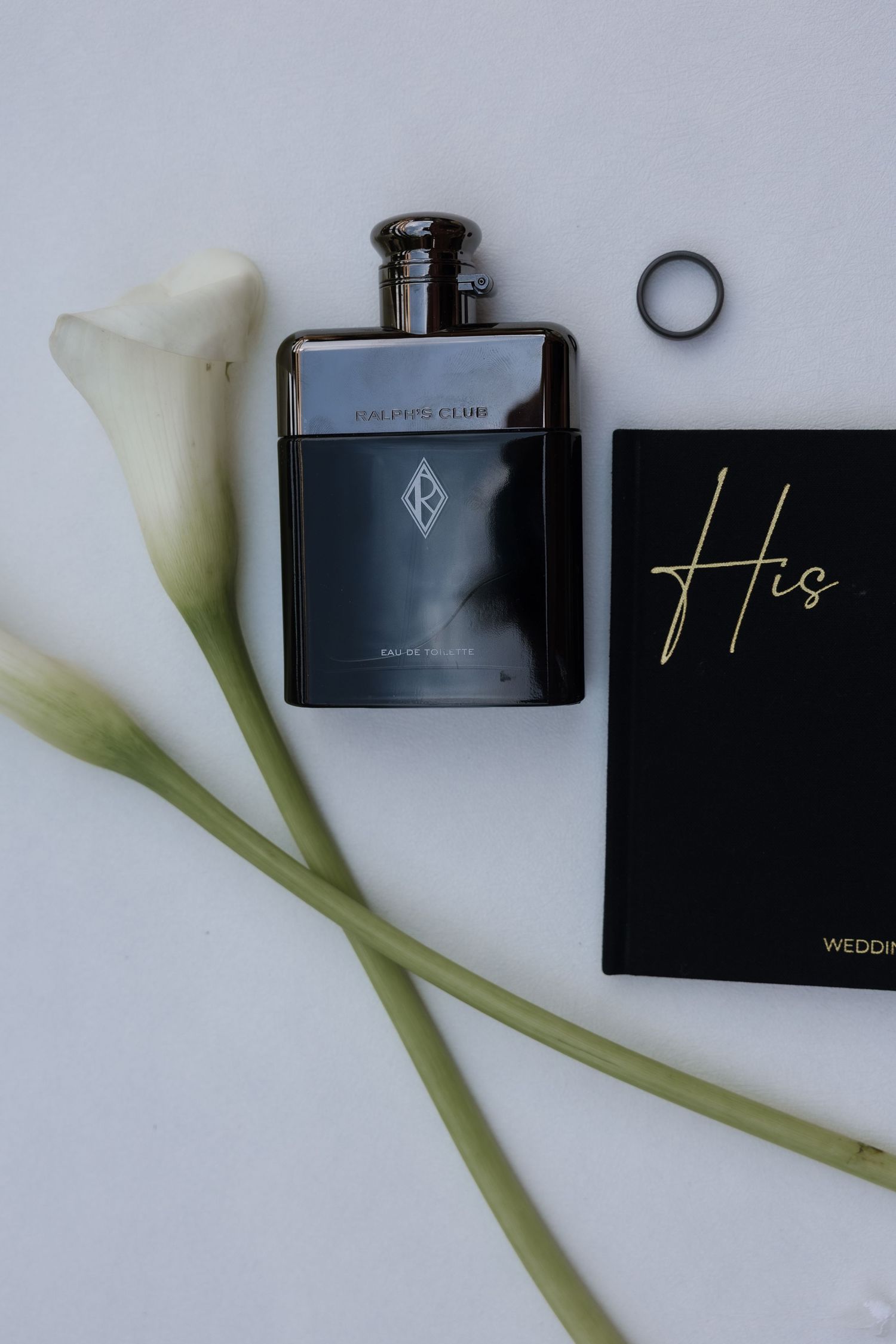 Groom wedding flat lay with dark cologne bottle, black ring, calla lilies, and His vow book.