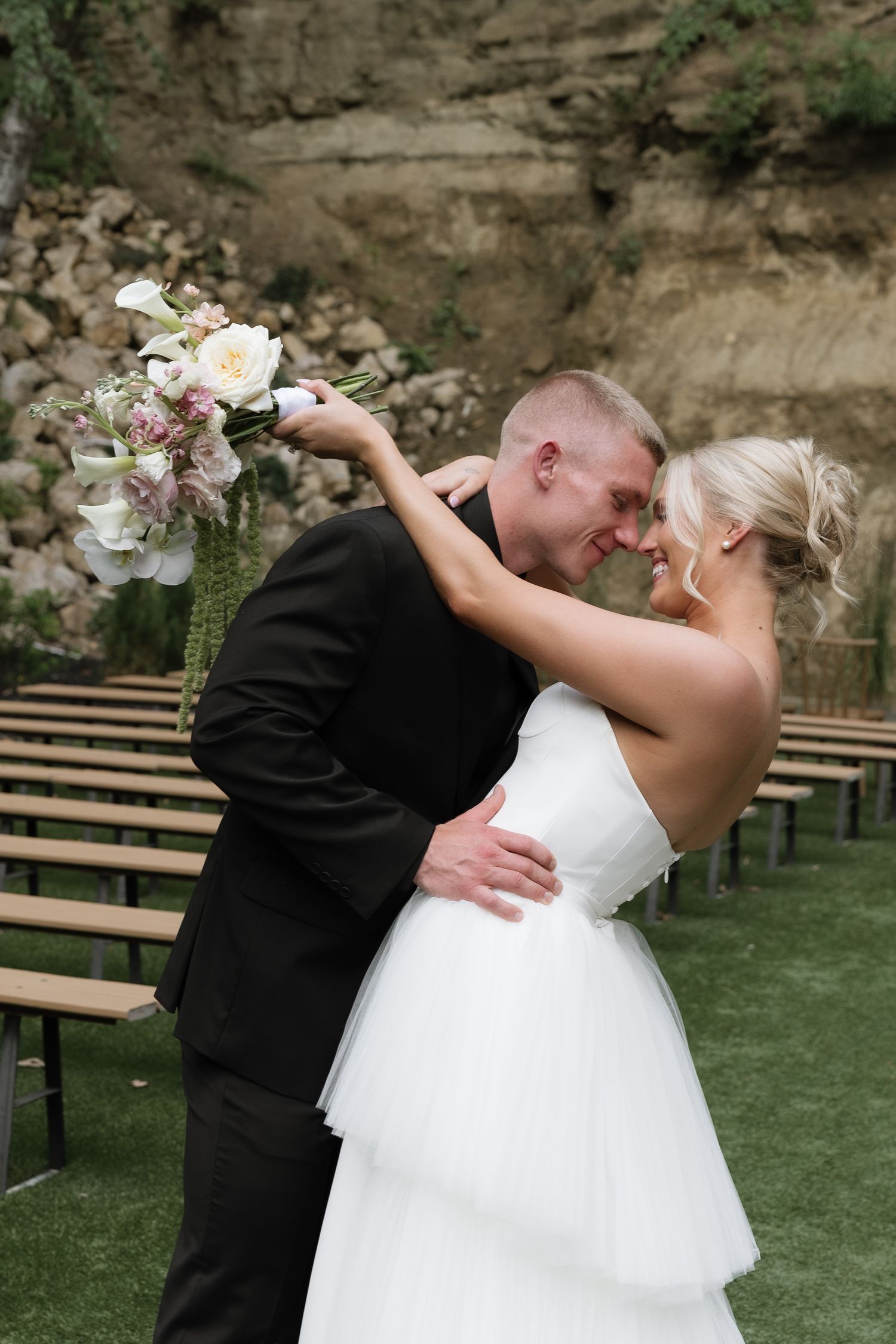 Bride and groom share a romantic moment, touching foreheads while bride holds a white floral bouquet at outdoor wedding venue.
