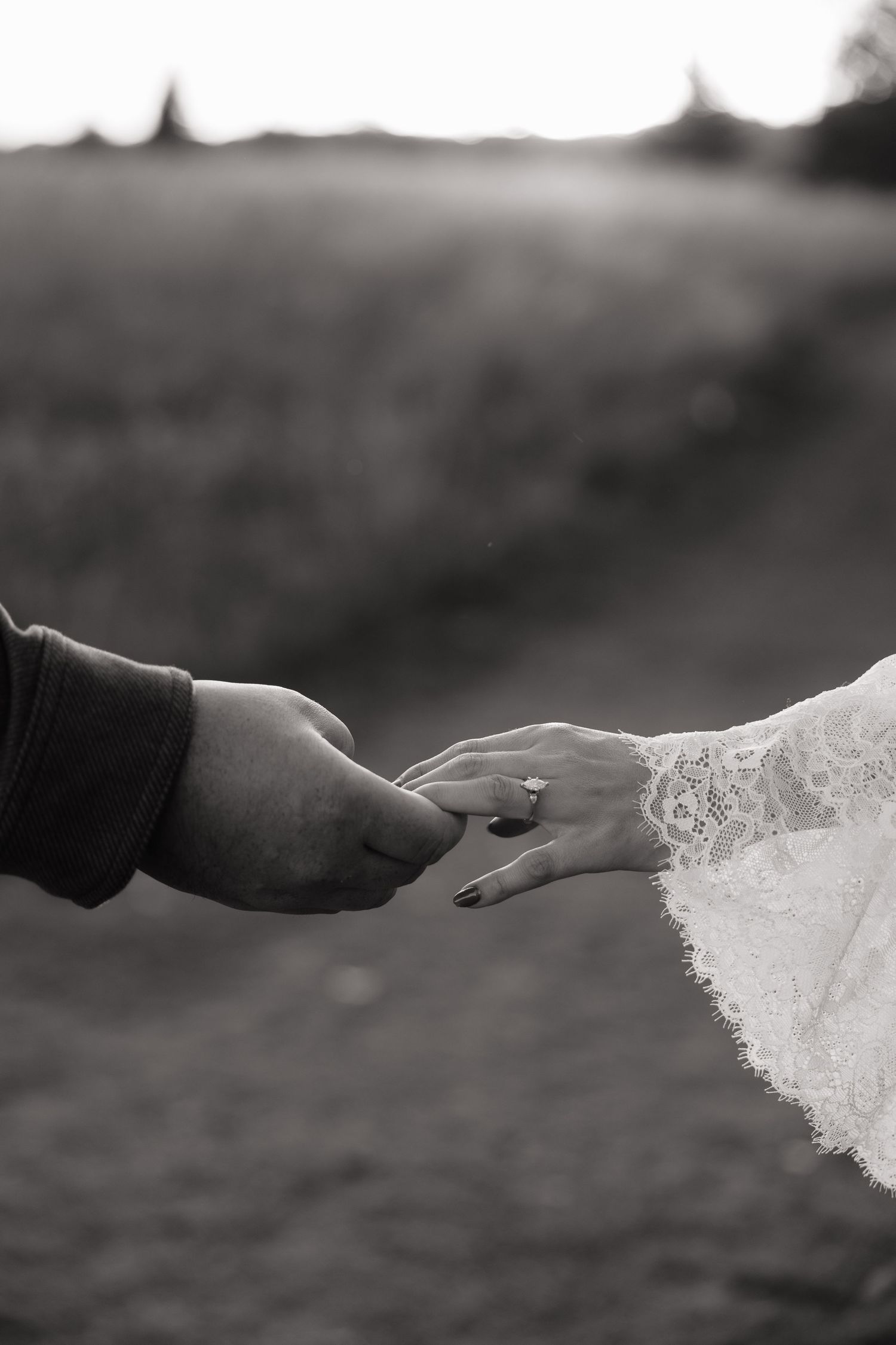 Black and white photo of bride and groom holding hands, wedding ring visible on brides finger, outdoor field backdrop.