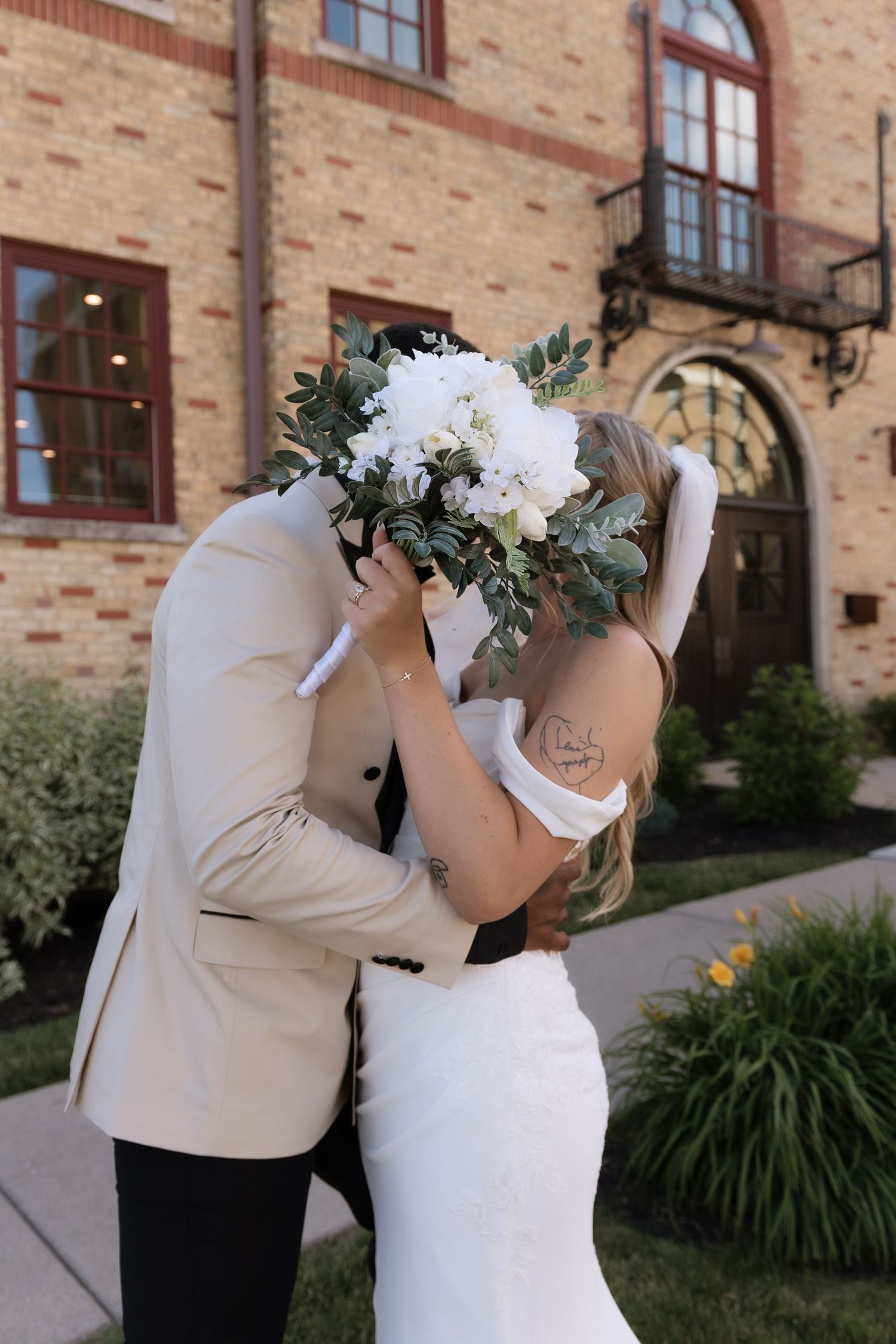 Bride and groom kissing behind white floral bouquet outside brick wedding venue.