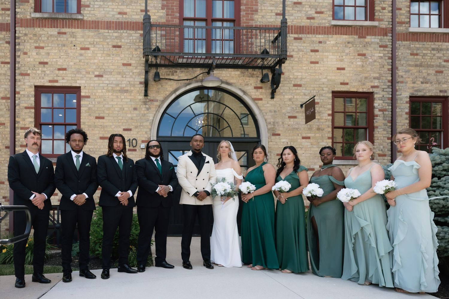 Wedding party poses outside brick venue, groomsmen in black suits, bridesmaids in sage green gowns, bride and groom centered.