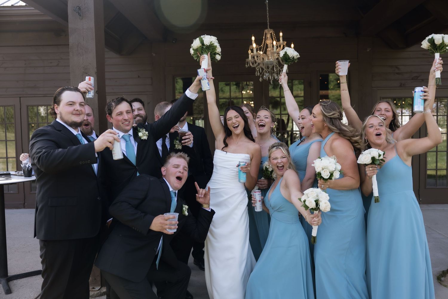 Excited wedding party in light blue bridesmaid dresses and suits celebrate together raising glasses and bouquets.