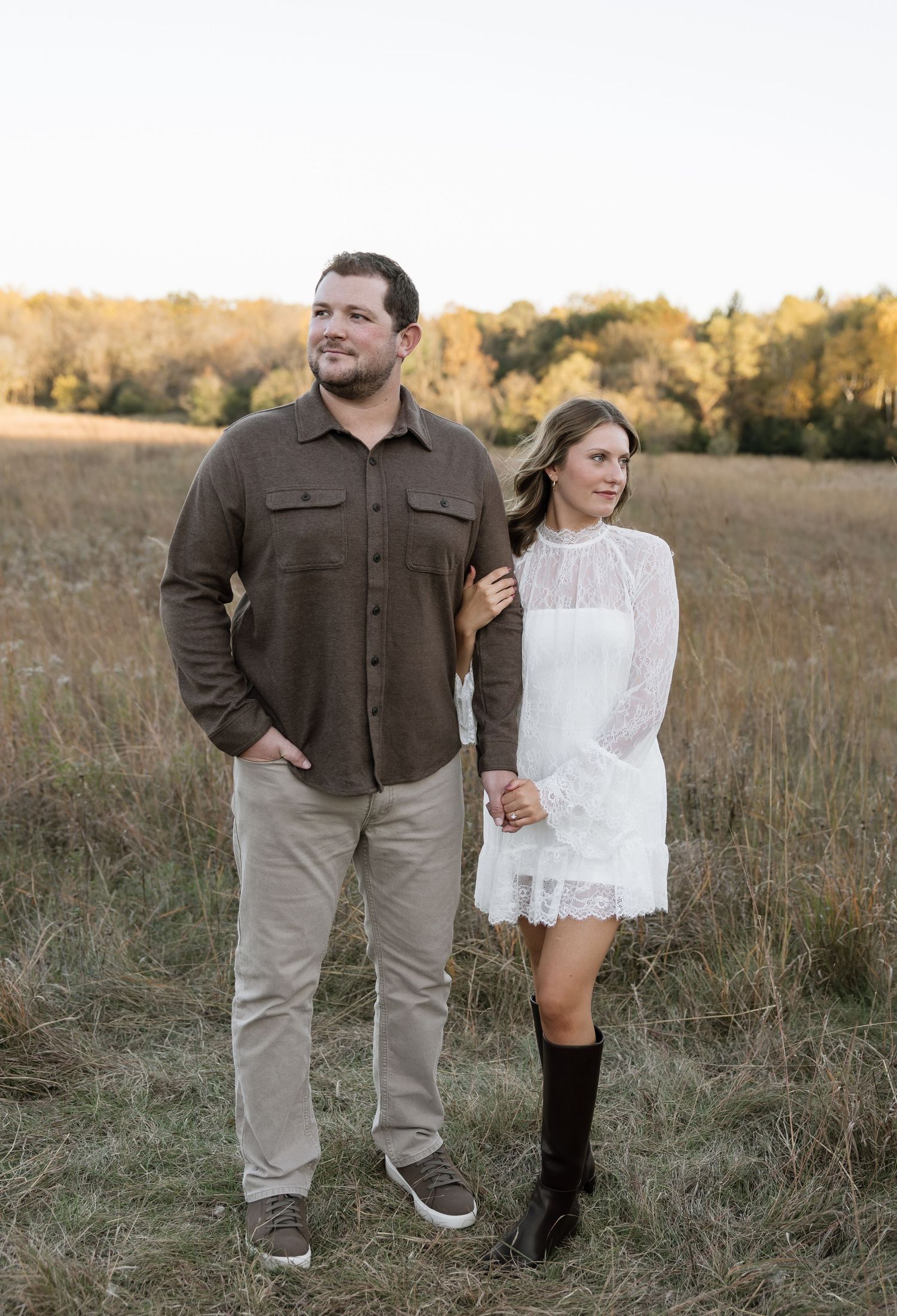 Couple holding hands in a golden field, man in brown shirt and khakis, woman in white lace dress with black boots.