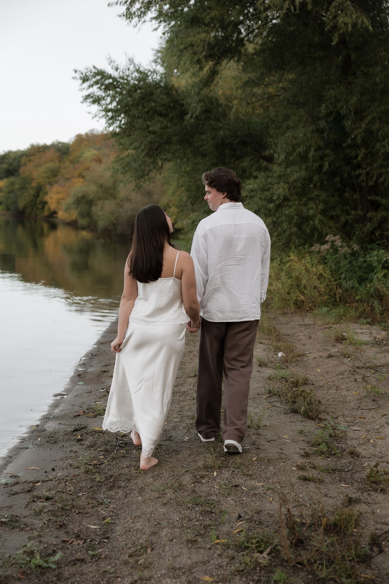 Engaged couple walking hand in hand along a sandy lakeside path surrounded by autumn foliage.