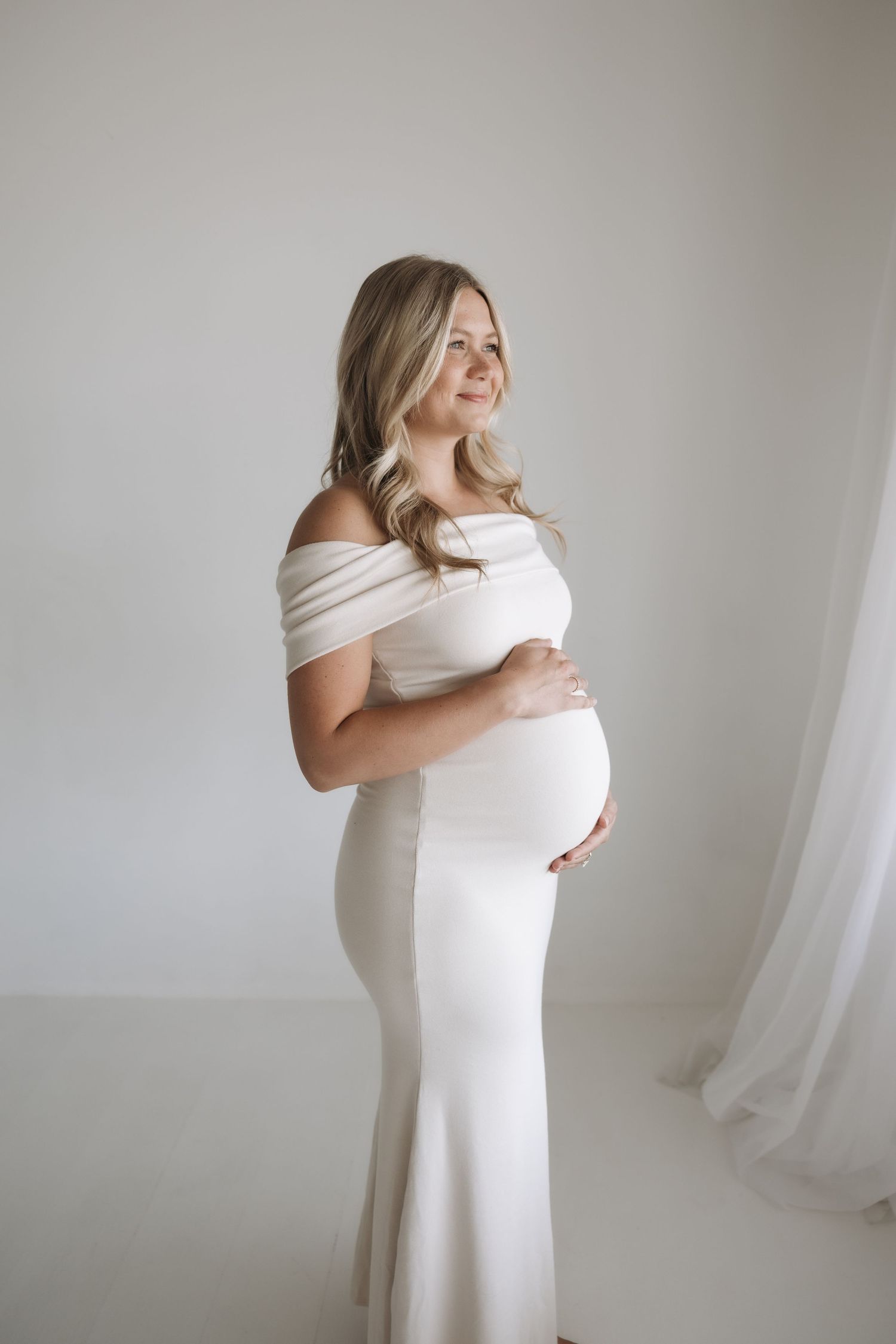 Smiling pregnant woman in off-shoulder white gown holding her baby bump in bright studio.
