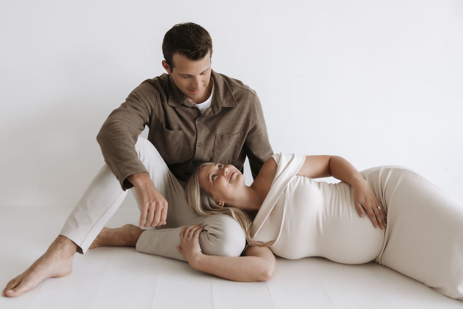 Pregnant woman in white dress resting her head on partner's lap on a white studio floor.