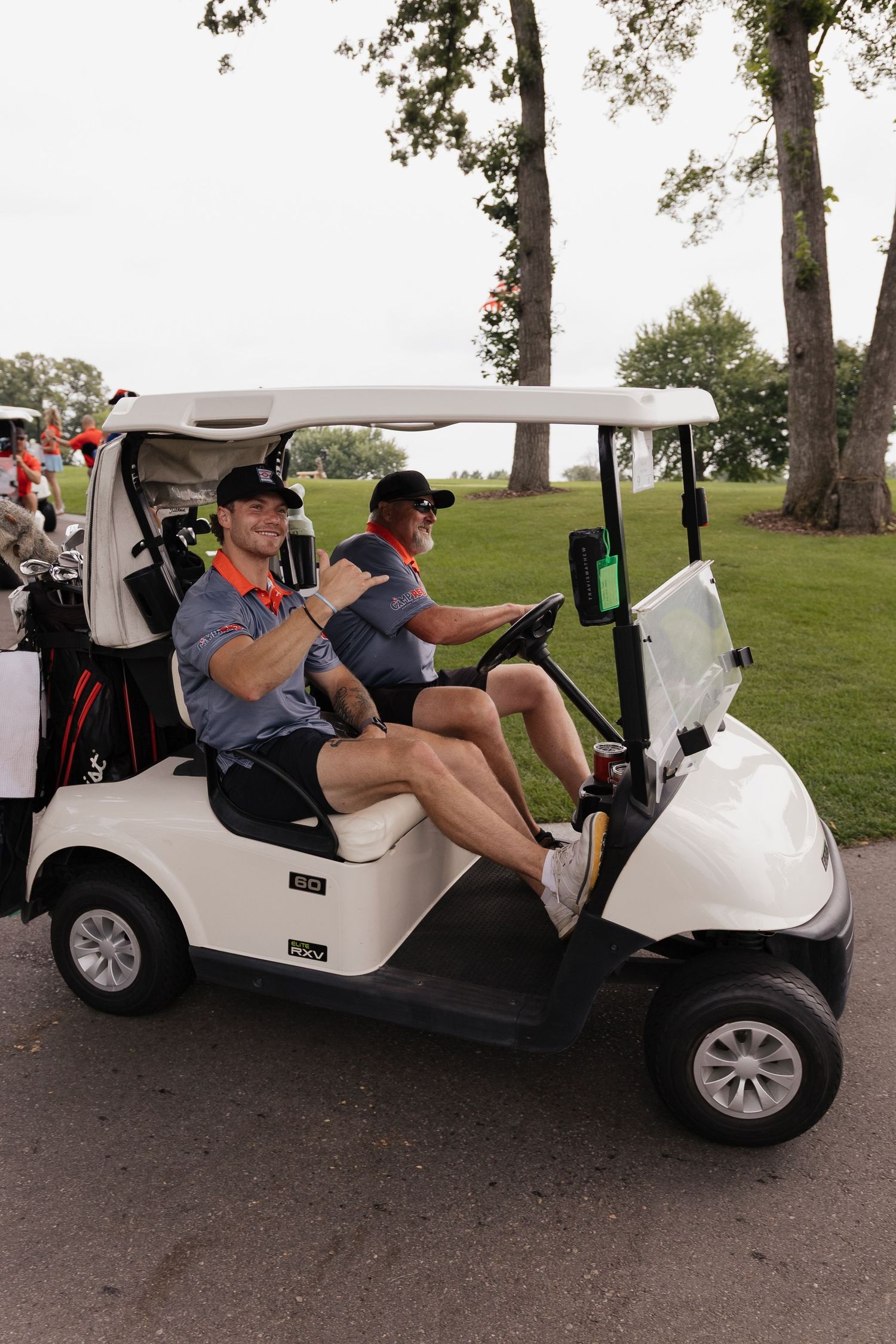 Two men smiling in a white golf cart on a golf course path surrounded by green trees.
