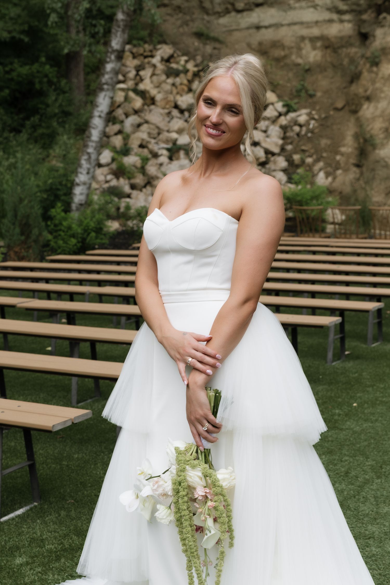 Beautiful bride in strapless layered white ballgown holding cascading bouquet at outdoor wedding venue.