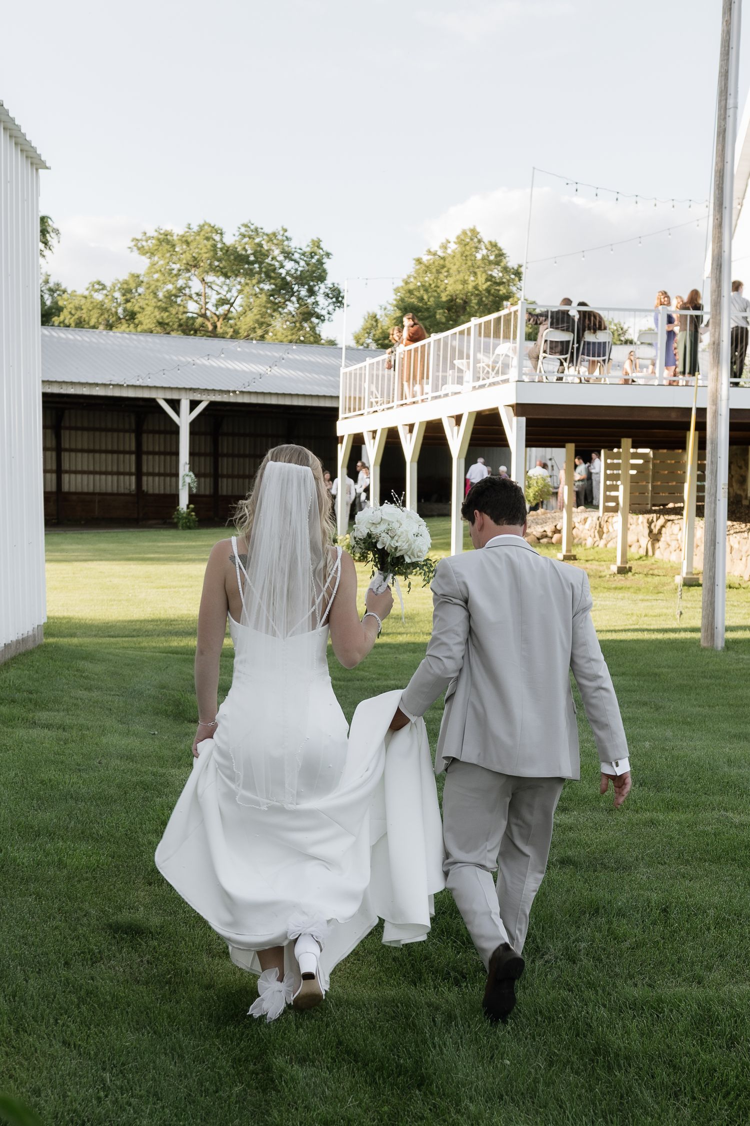 Bride and groom walk hand-in-hand across green lawn toward decorated deck at outdoor barn wedding venue.