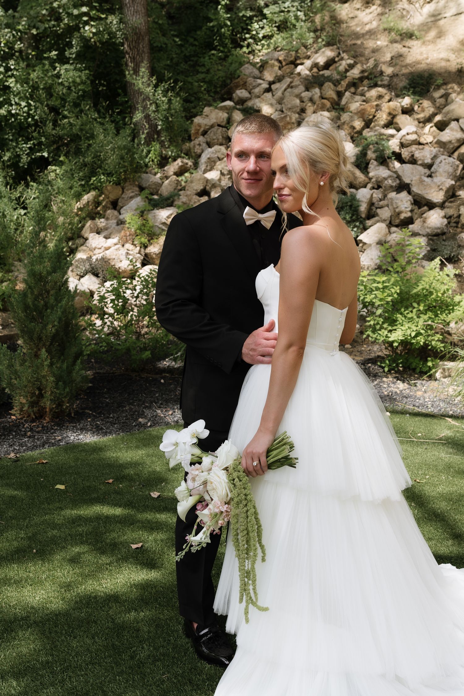 Smiling bride and groom pose in garden setting, she wears strapless tiered gown holding white and green bouquet.