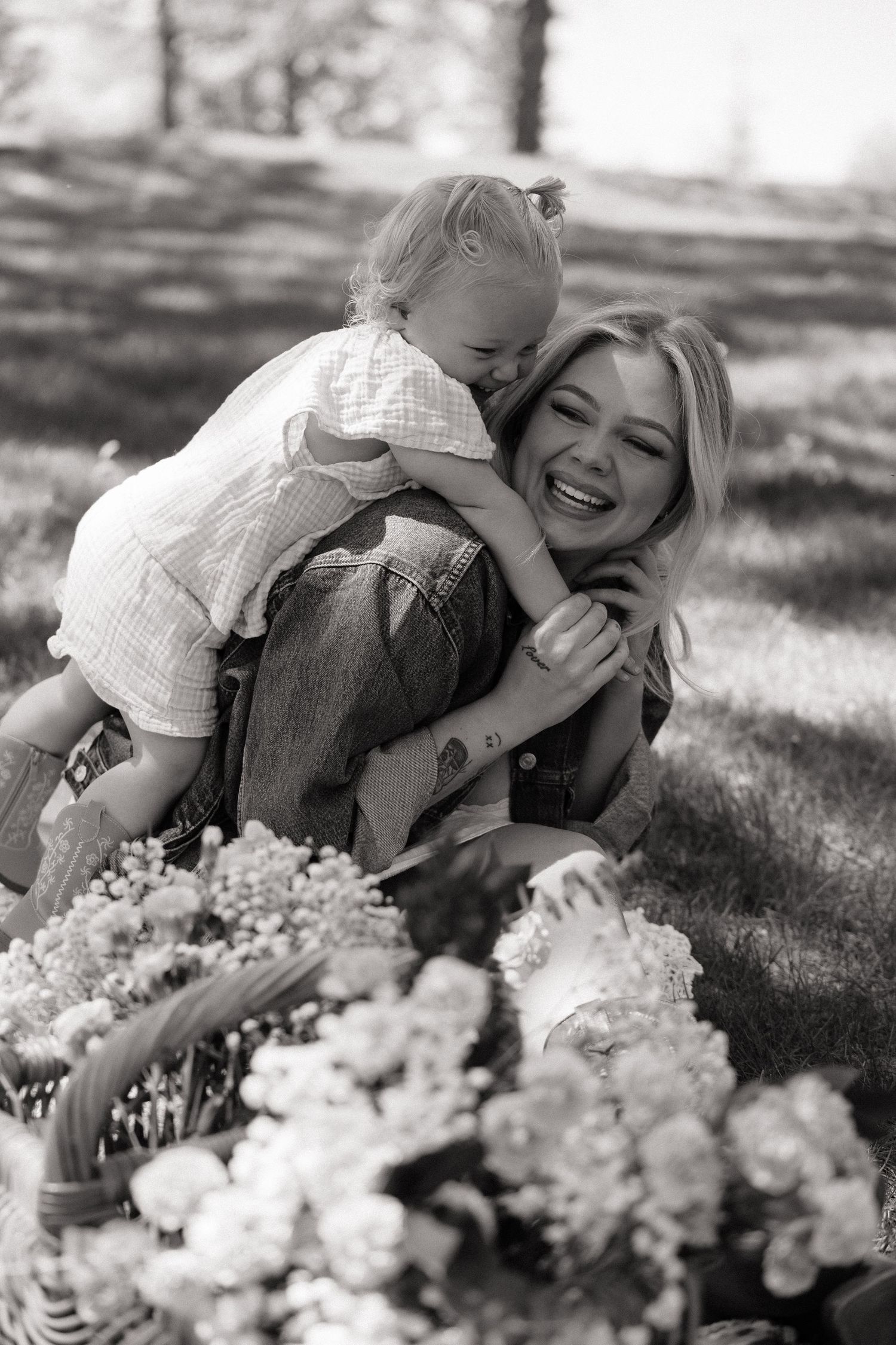 Black and white photo of laughing mother being hugged by her joyful toddler daughter outdoors.