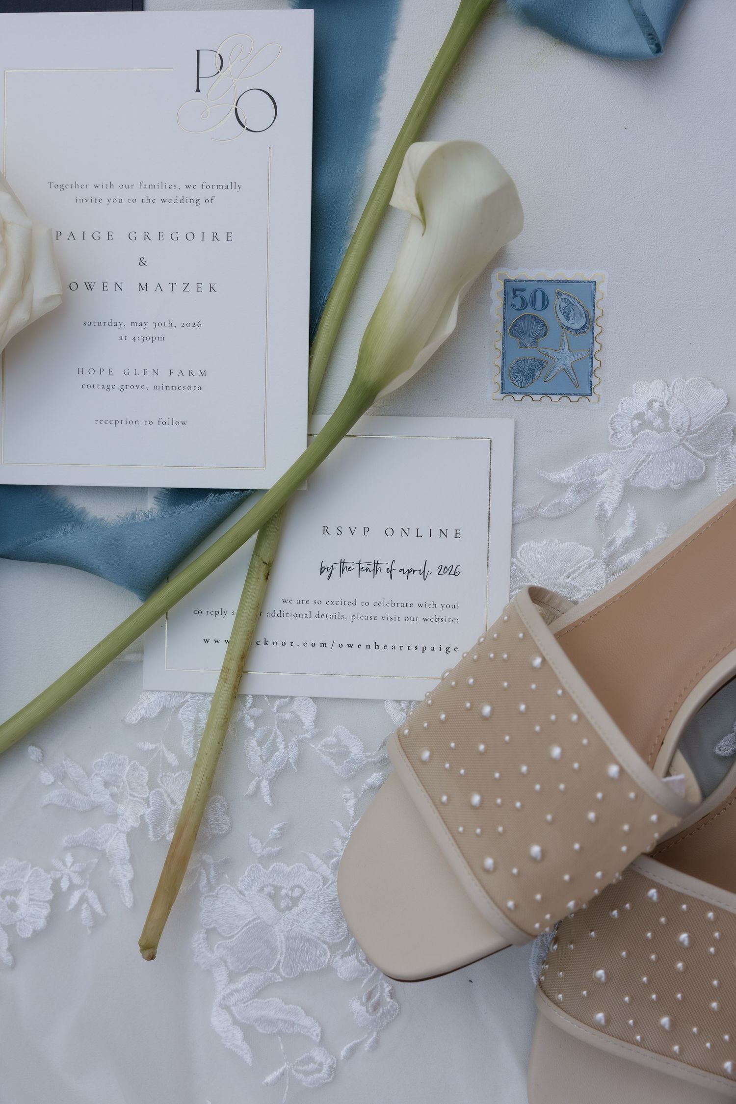 Wedding flat lay with invitation, pearl heels, calla lily, stamps, and blue ribbon.