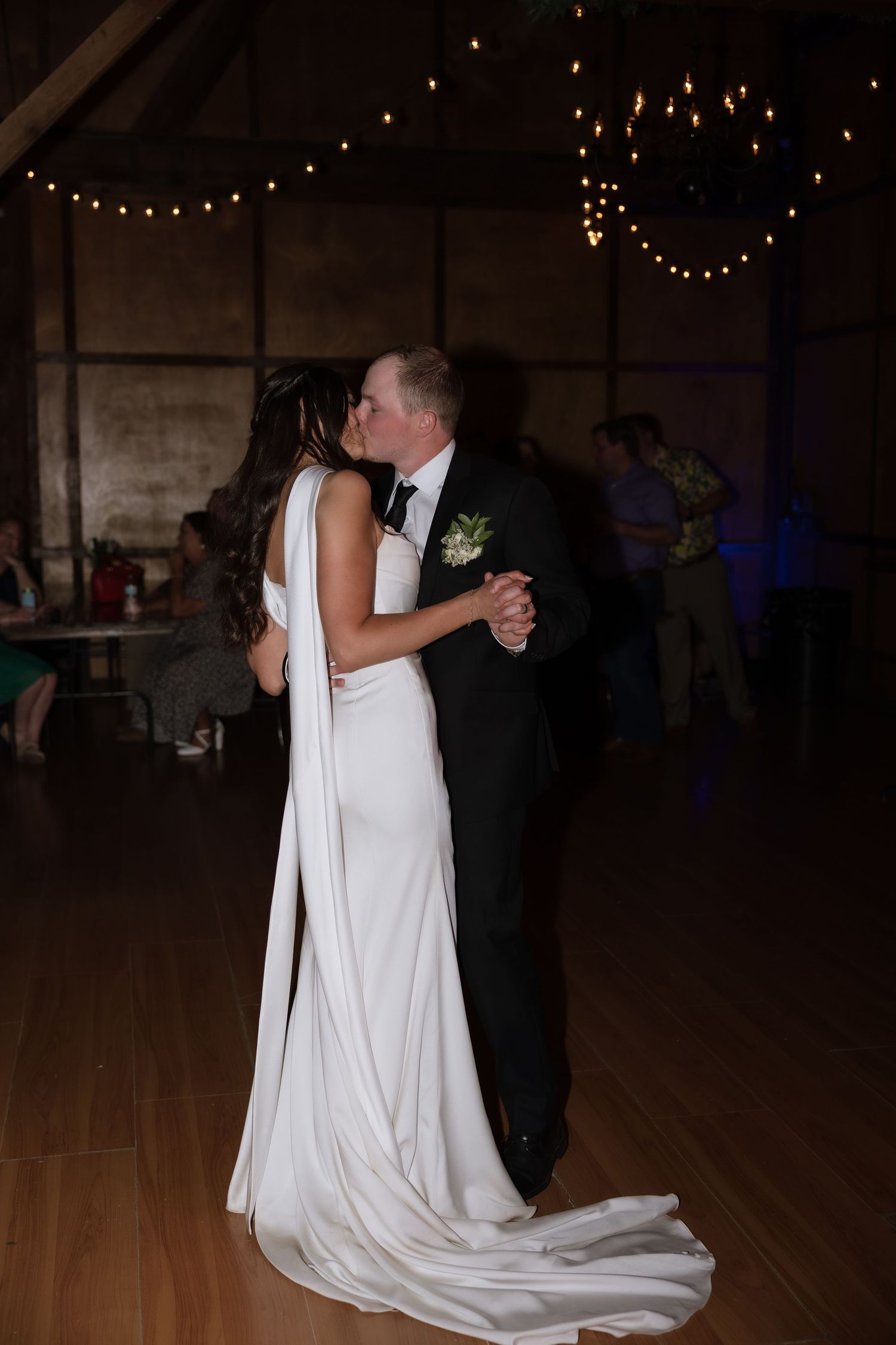 Bride and groom share their first dance kiss under warm string lights in a rustic barn reception.