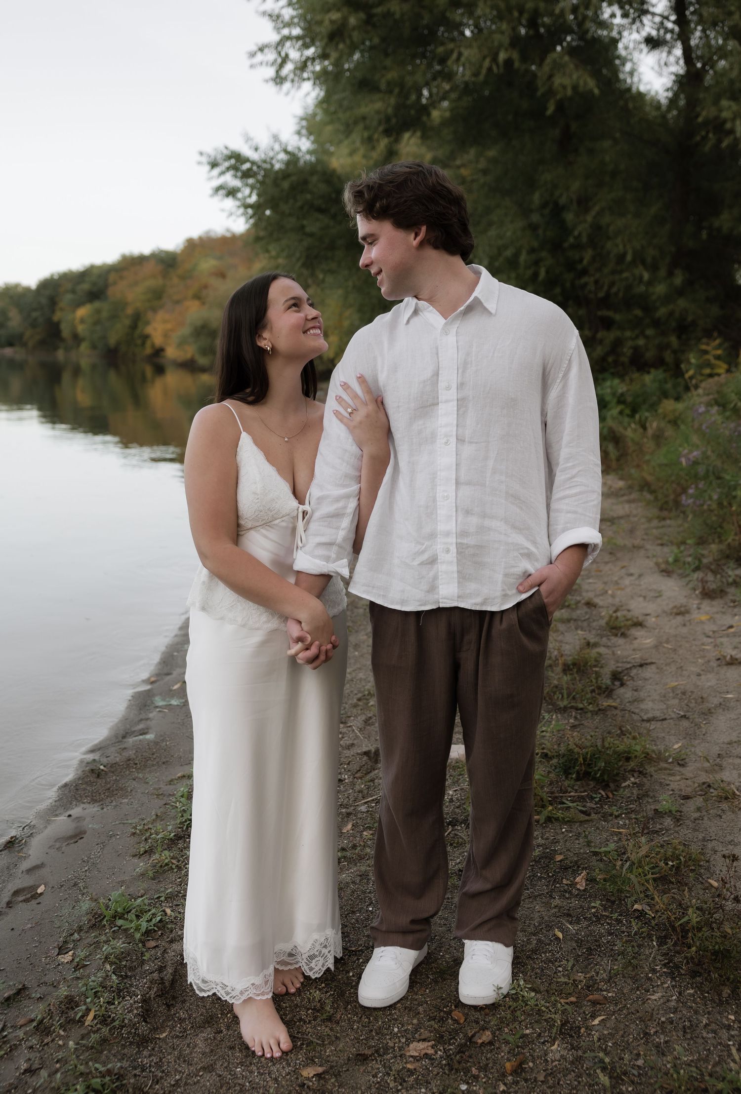Happy couple standing by a lakeside, woman in white slip dress looking up at man in white linen shirt.