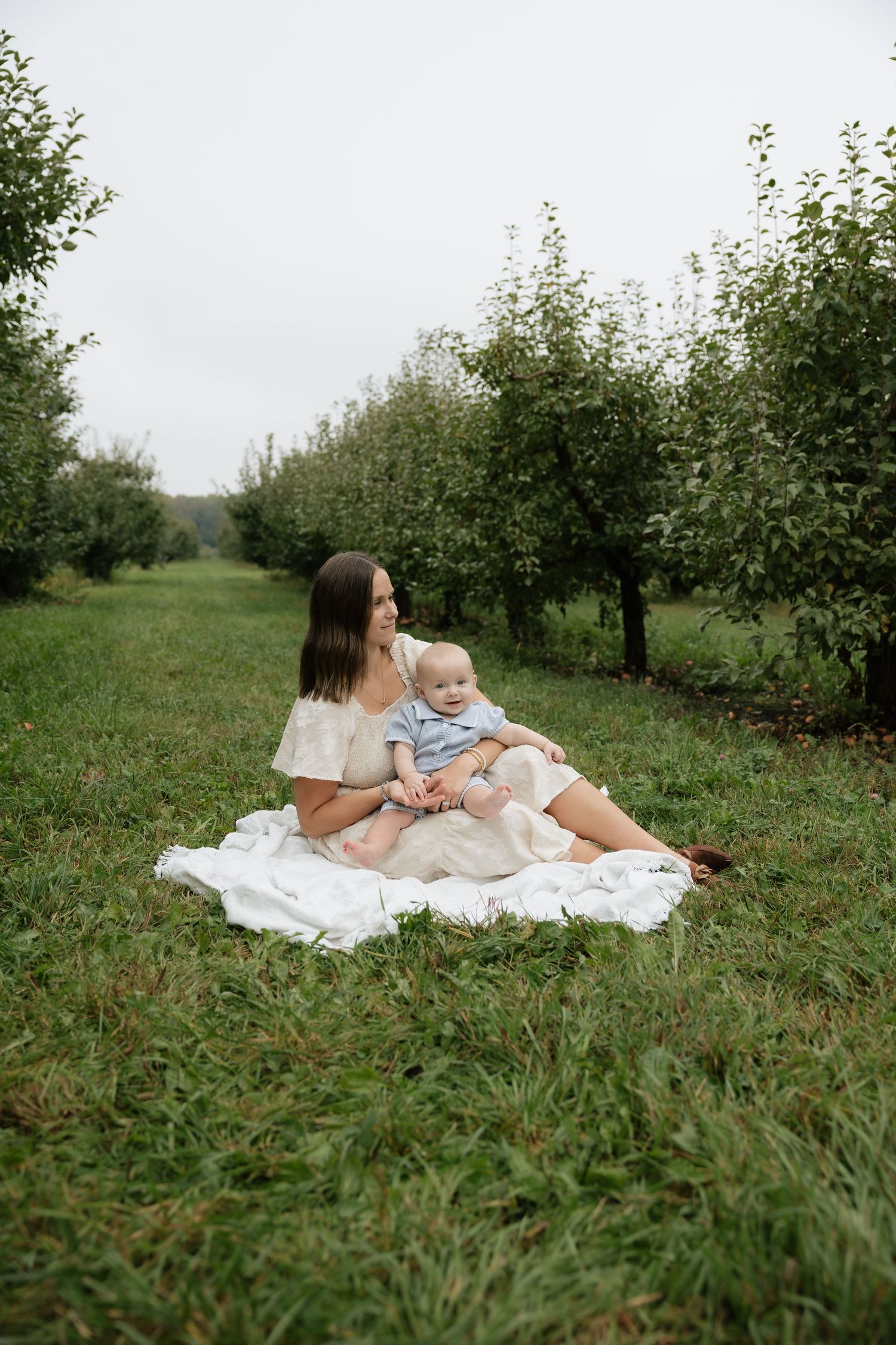 Mother sitting on white blanket in apple orchard holding baby in blue outfit surrounded by green grass and trees.