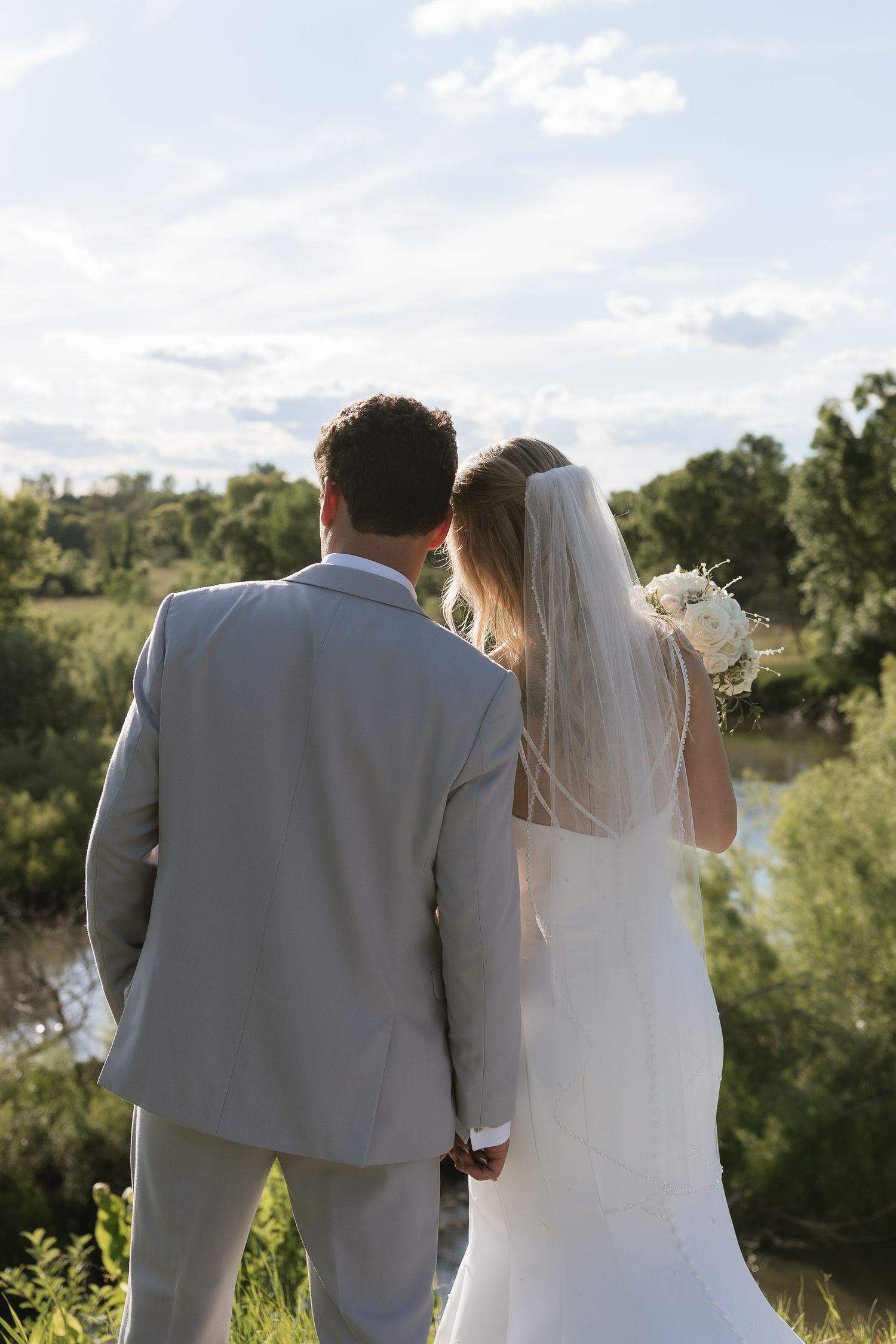 A bride and groom stand hand in hand overlooking a scenic river landscape on their wedding day.
