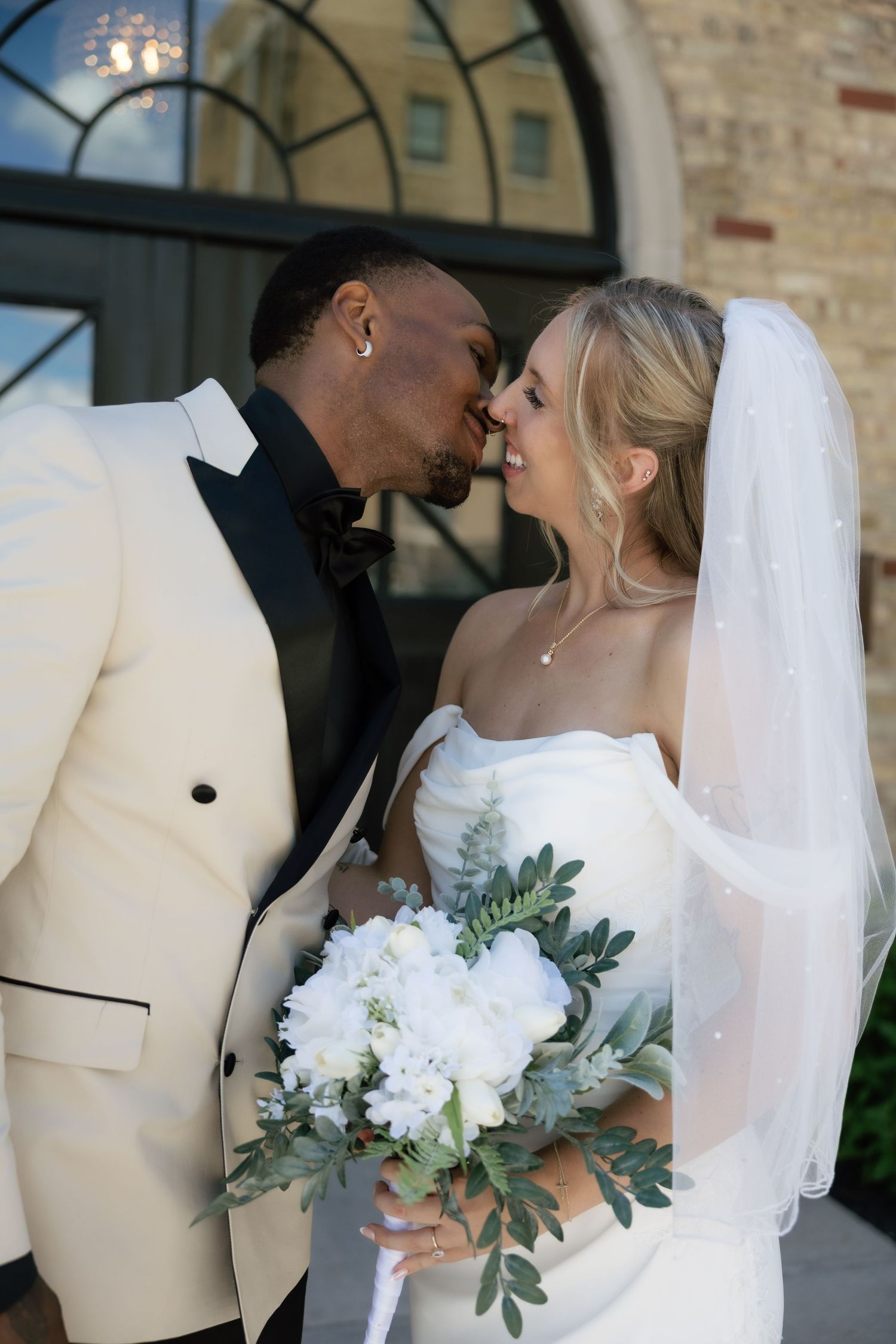 Interracial bride and groom nearly kissing outdoors, bride in strapless gown with veil holding white floral bouquet.
