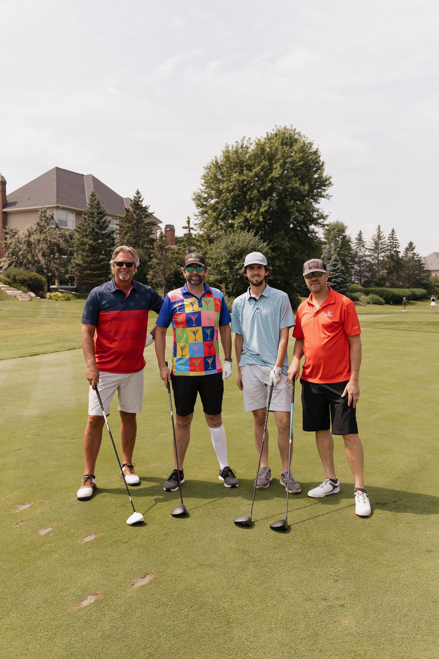 Four men holding golf clubs pose together on a sunny golf course green, wearing colorful summer attire.
