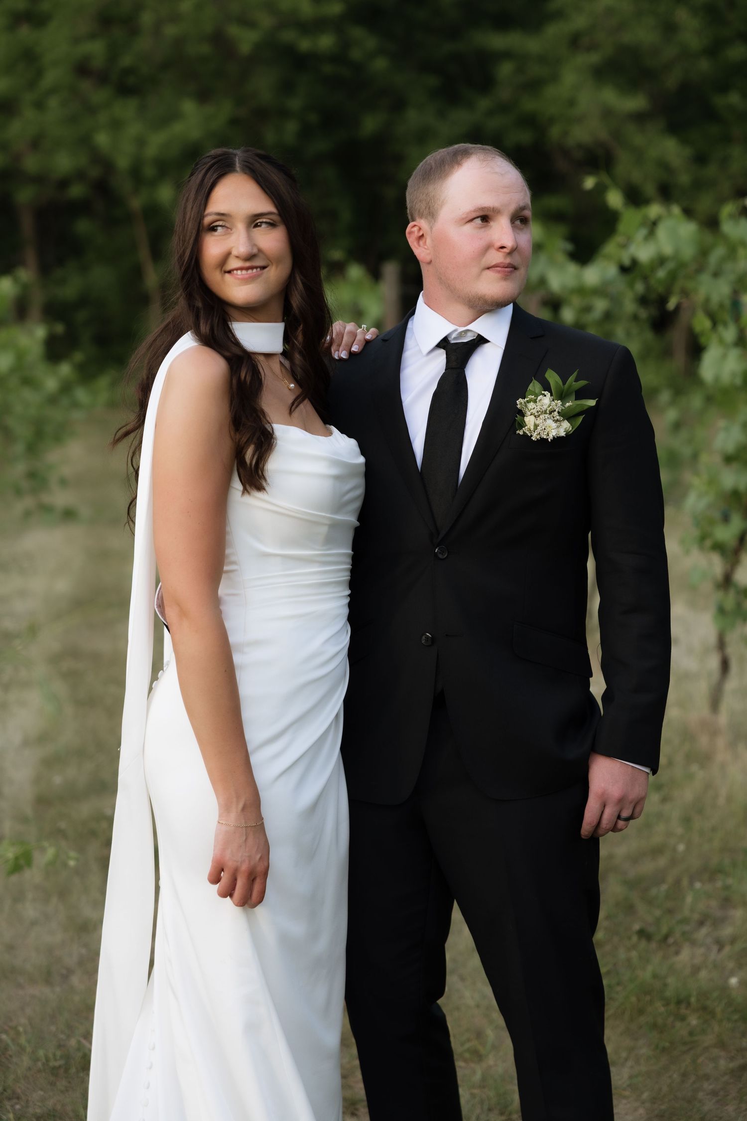 Bride in a white halter gown and groom in a black suit pose together outdoors surrounded by lush greenery.
