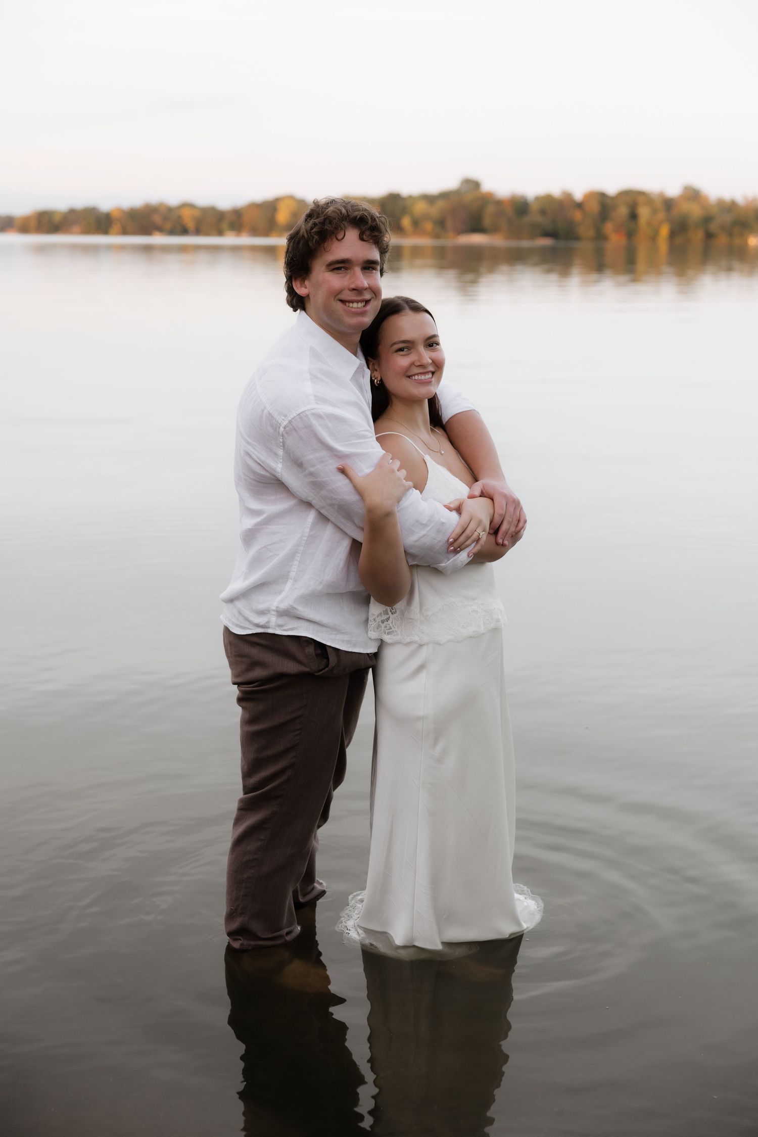 Couple standing in lake water, man in white shirt embracing woman in white dress, trees reflected in calm water.