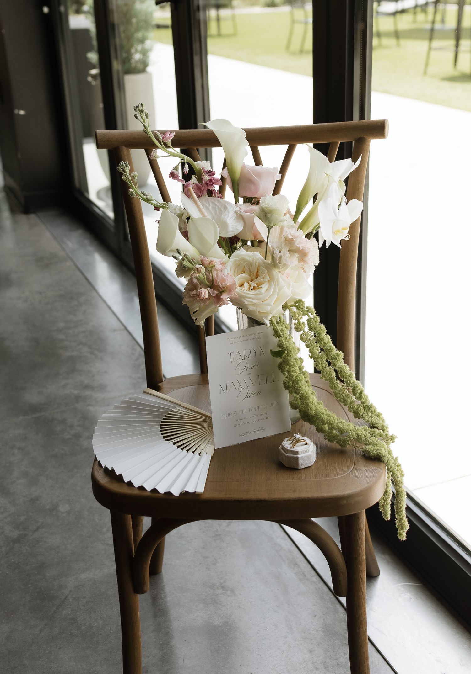 Elegant wedding chair with floral bouquet, paper fan, and stationery displayed near large windows.