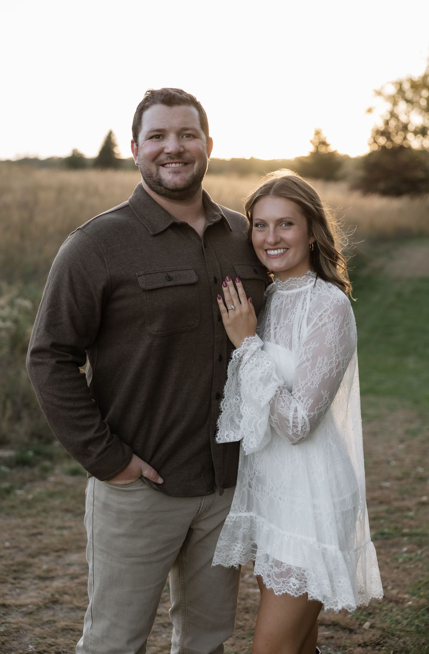 Smiling couple posing outdoors at golden hour, woman in white lace dress and man in brown shirt.