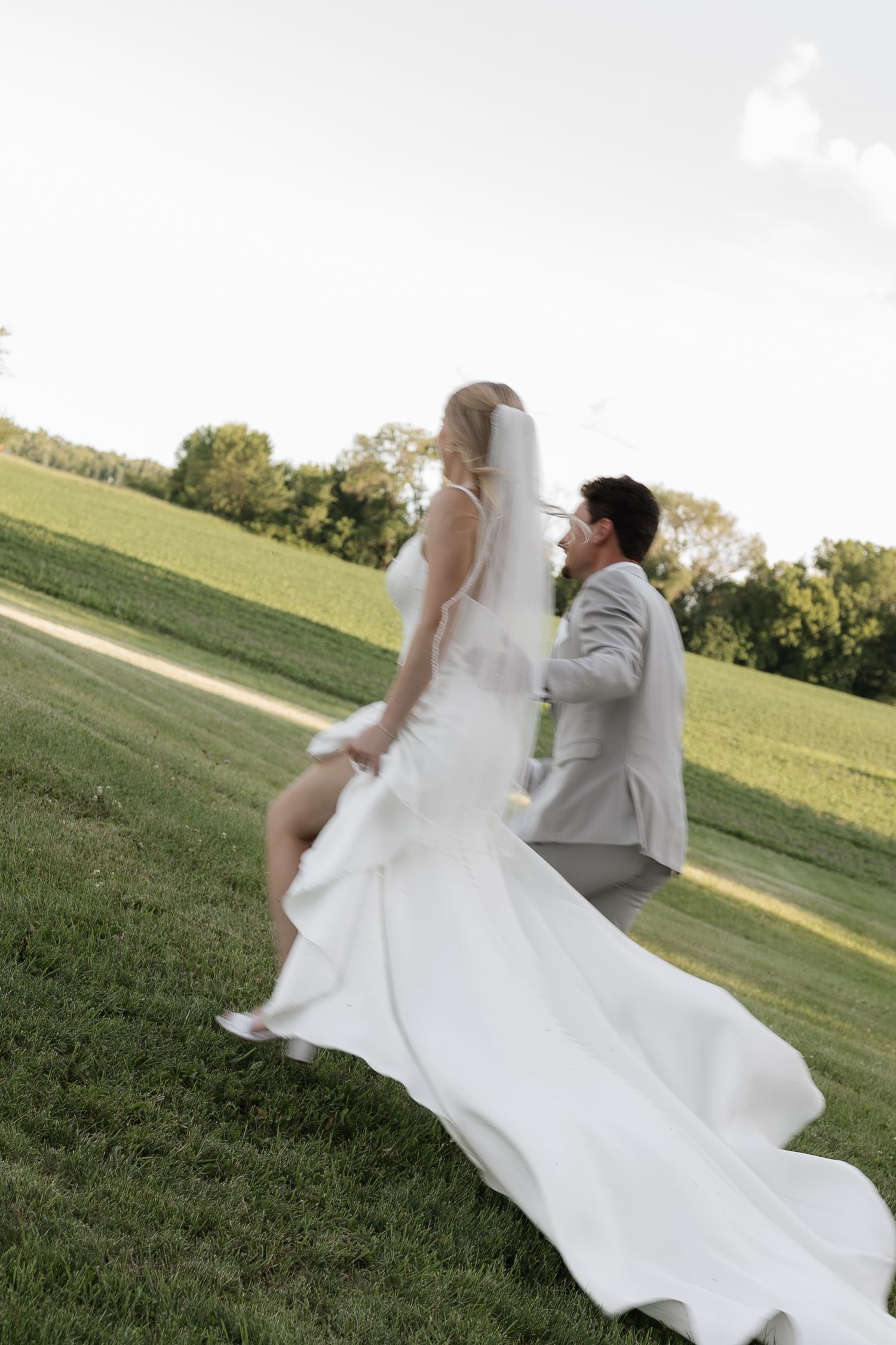 Bride in white gown and veil walks with groom in gray suit across lush green lawn at outdoor wedding.