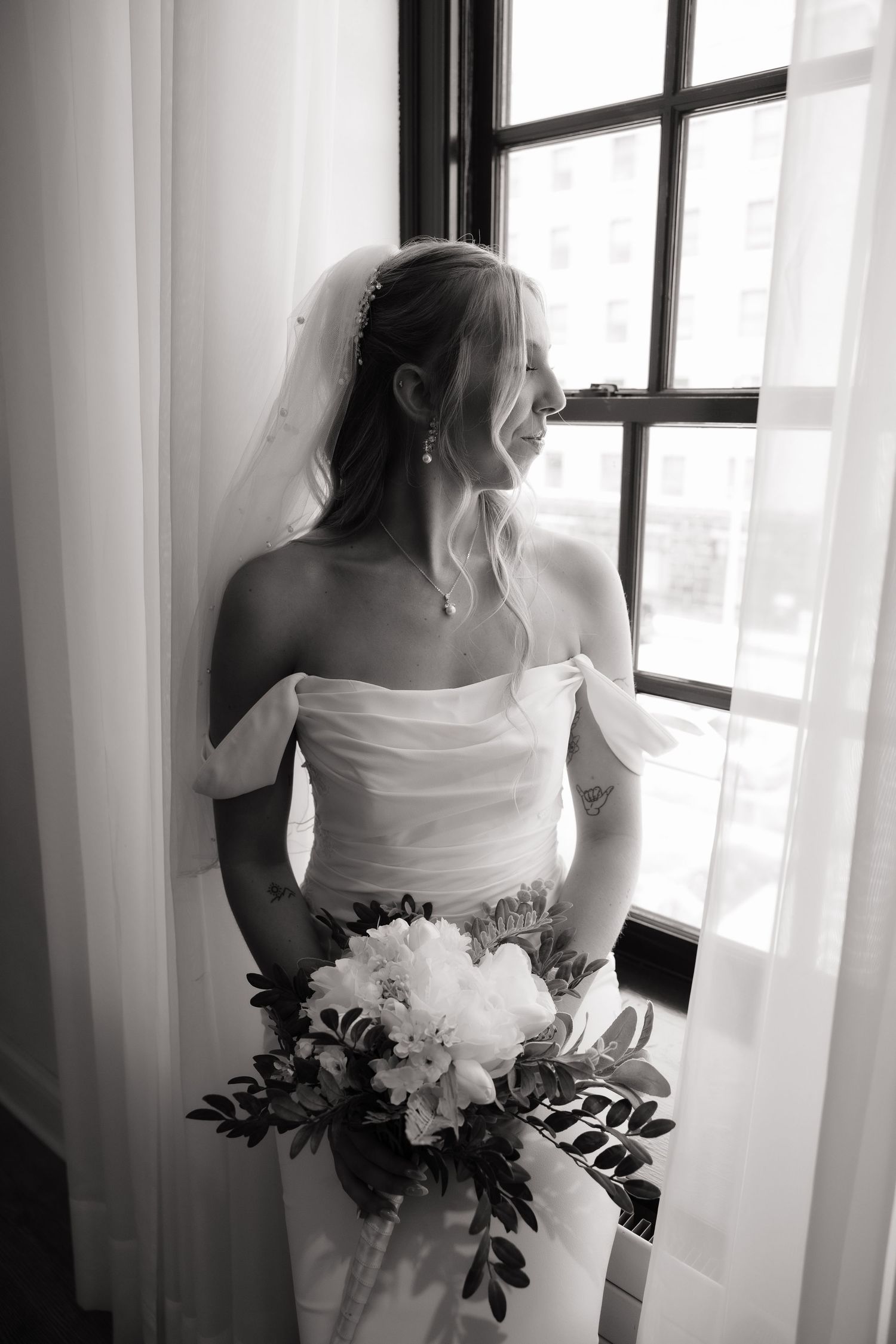 Black and white photo of a smiling bride in an off-shoulder gown holding a bouquet, standing by a window in soft light.