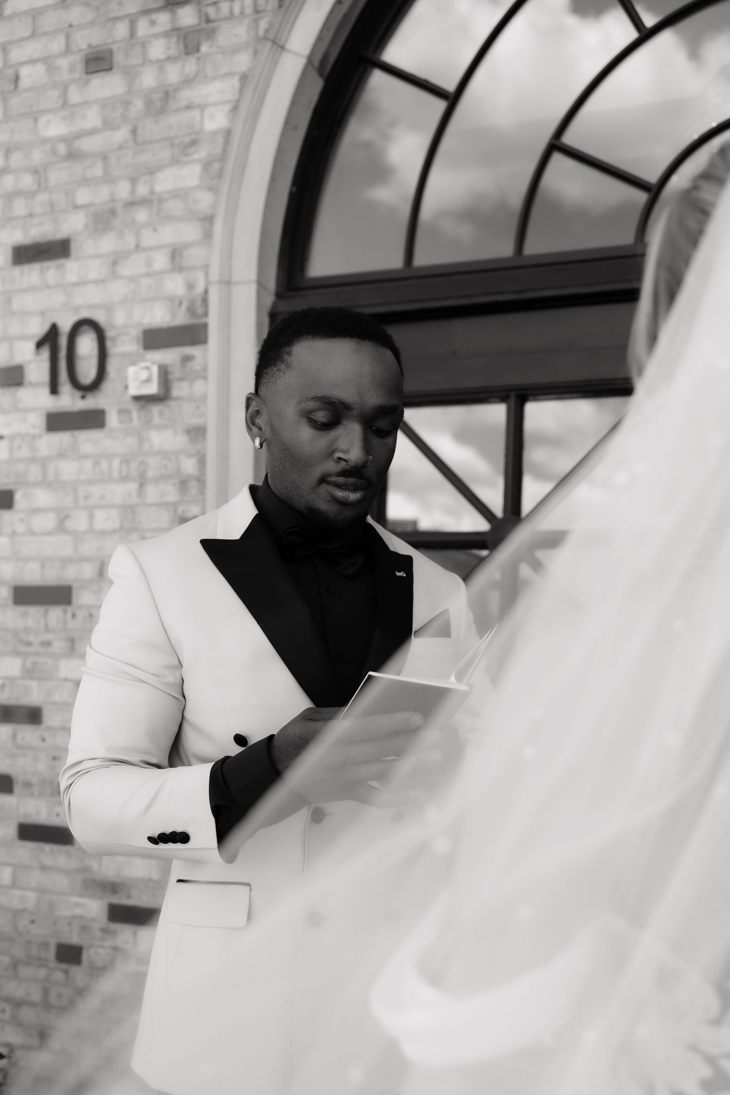 Black and white photo of a groom in a white tuxedo reading vows outside a brick building with an arched window.
