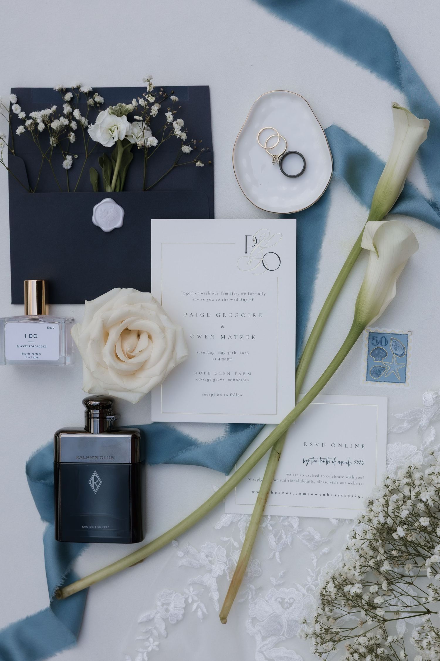 Elegant wedding flat lay with invitation suite, perfume, rings, calla lilies, and blue ribbon.