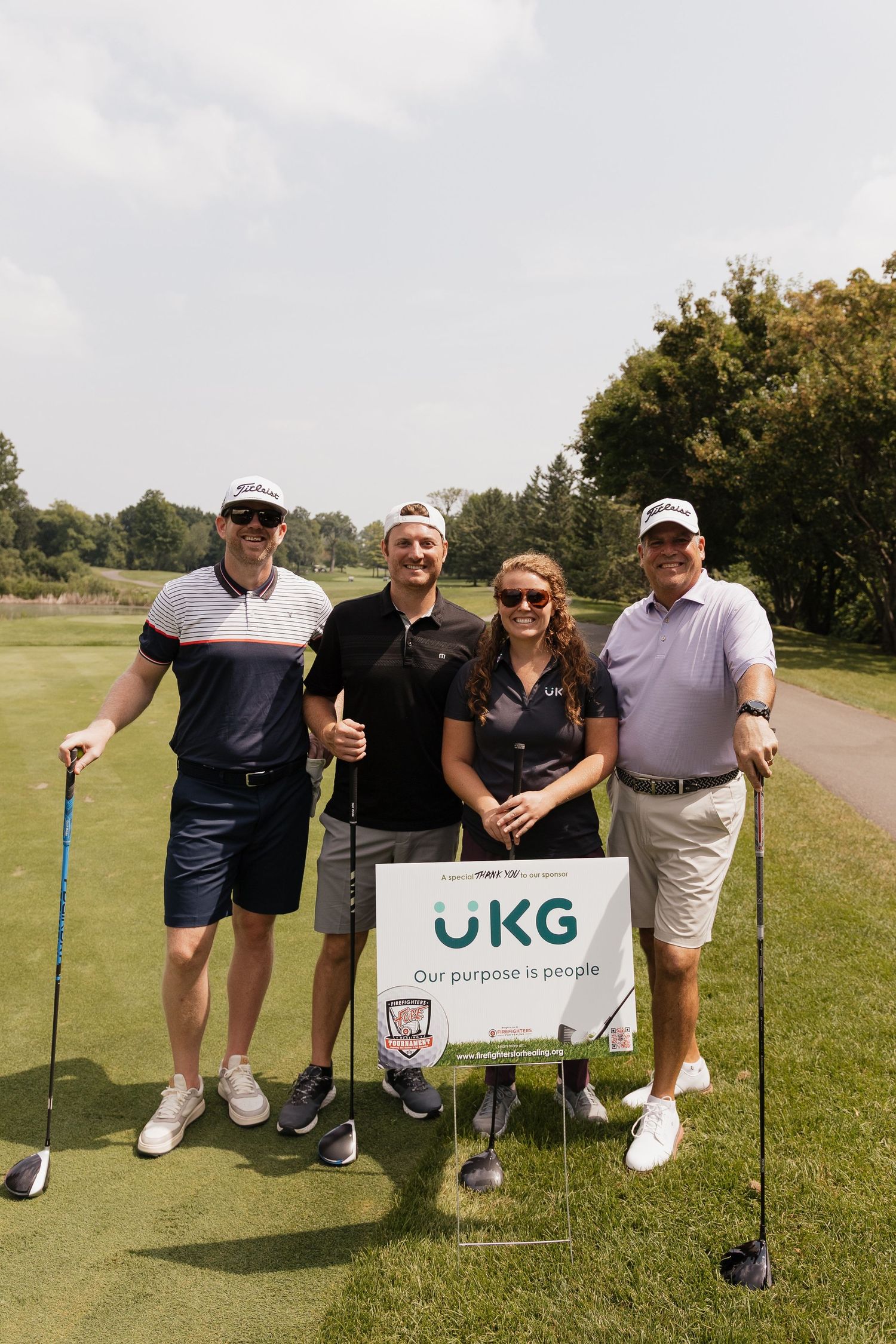Four golfers pose with a UKG 'Our purpose is people' sign on a sunny golf course.