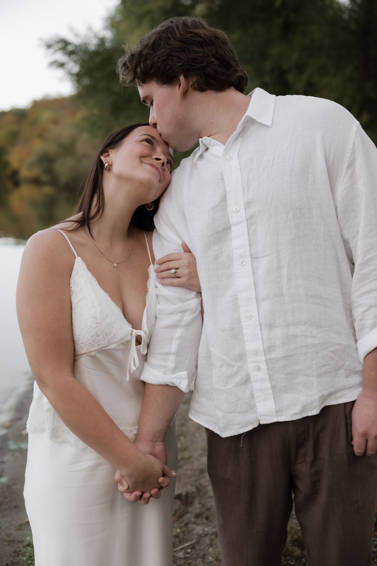 A couple shares a tender moment by a lakeside, the man kissing the woman's temple as she smiles in a white dress.