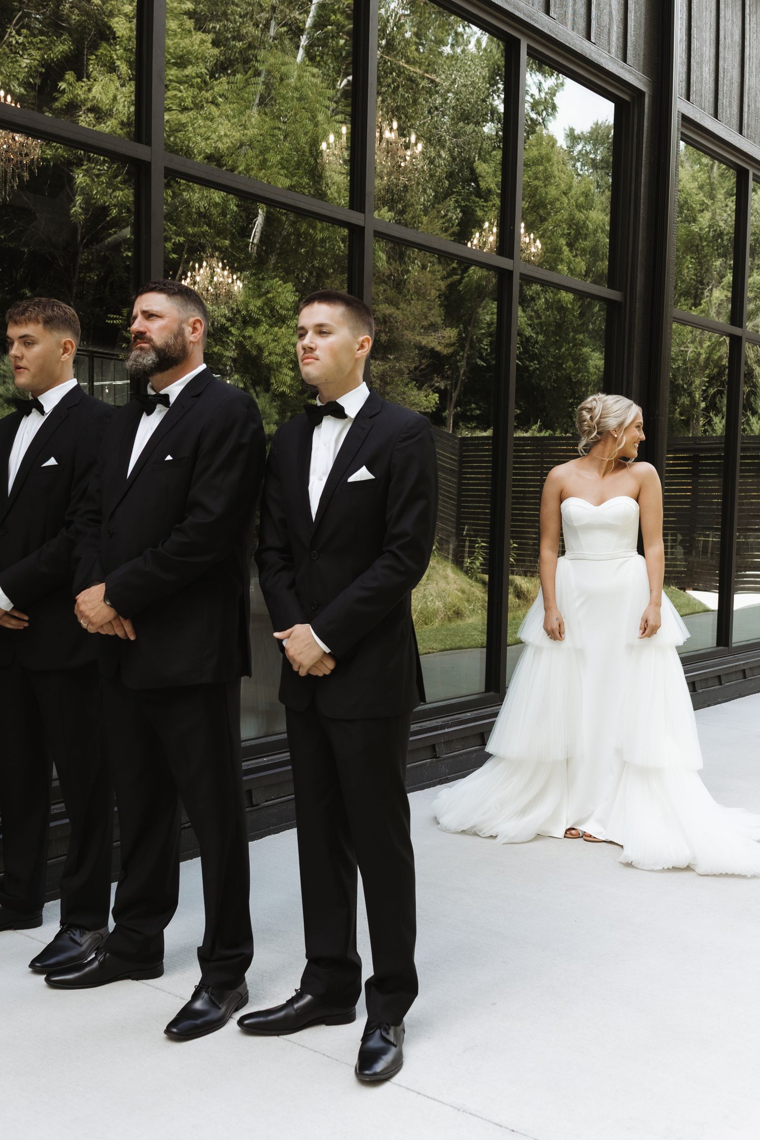 Groomsmen in black tuxedos stand at attention as bride in strapless tiered gown approaches ceremony.