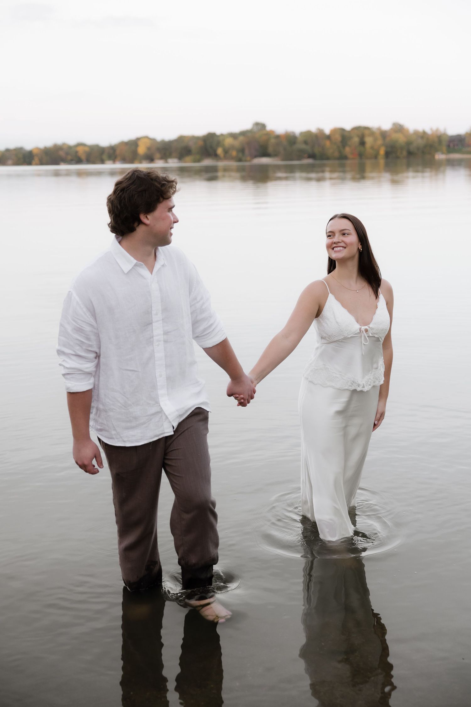 Smiling engaged couple holding hands while wading together in a calm lake with autumn trees behind.