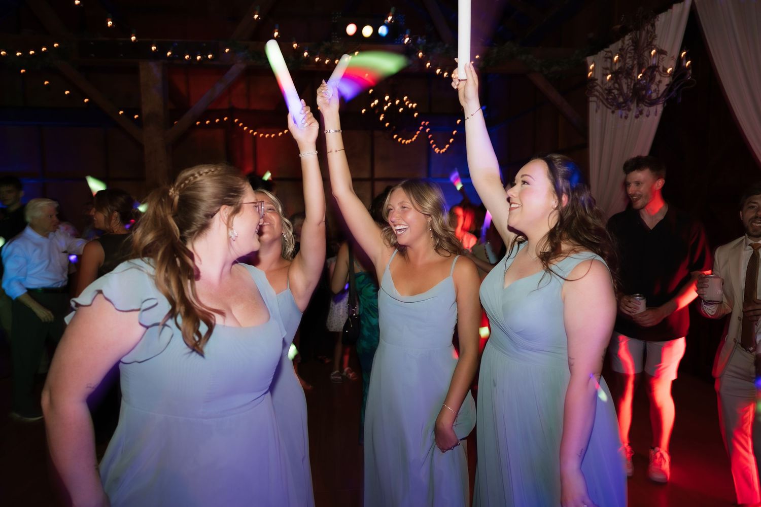 Bridesmaids in light blue gowns dance joyfully with glow sticks at wedding reception under string lights.
