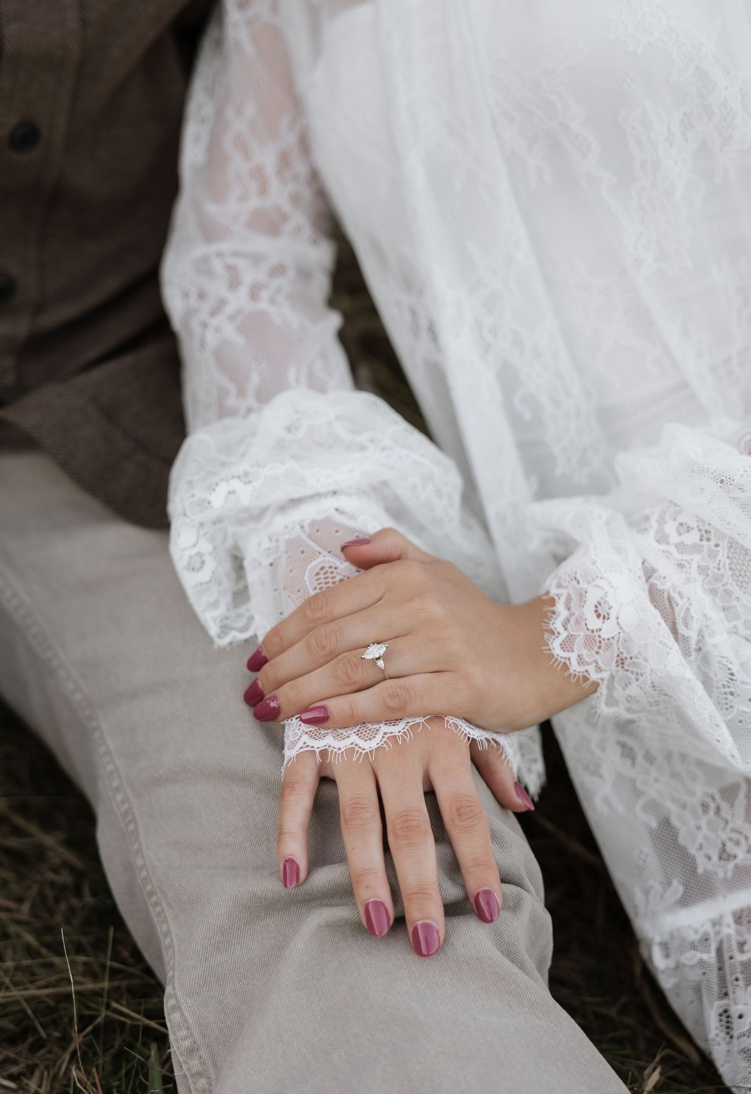 Close-up of a bride's hands in white lace dress sleeves, wearing a diamond engagement ring with mauve nails.